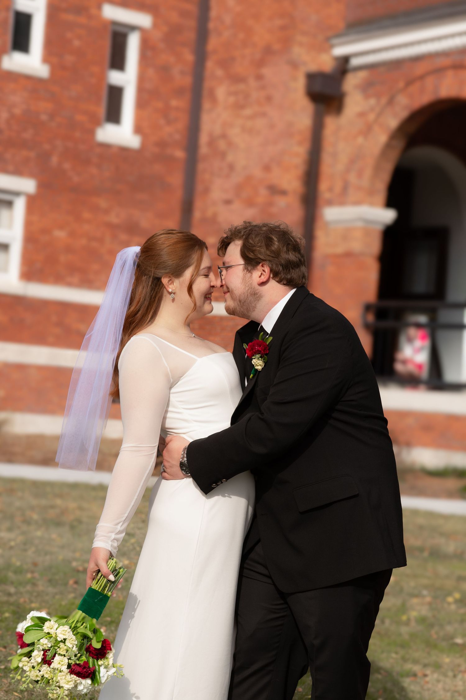 Bride and groom share romantic moment, nearly kissing, outside red brick venue, bride holding bouquet.