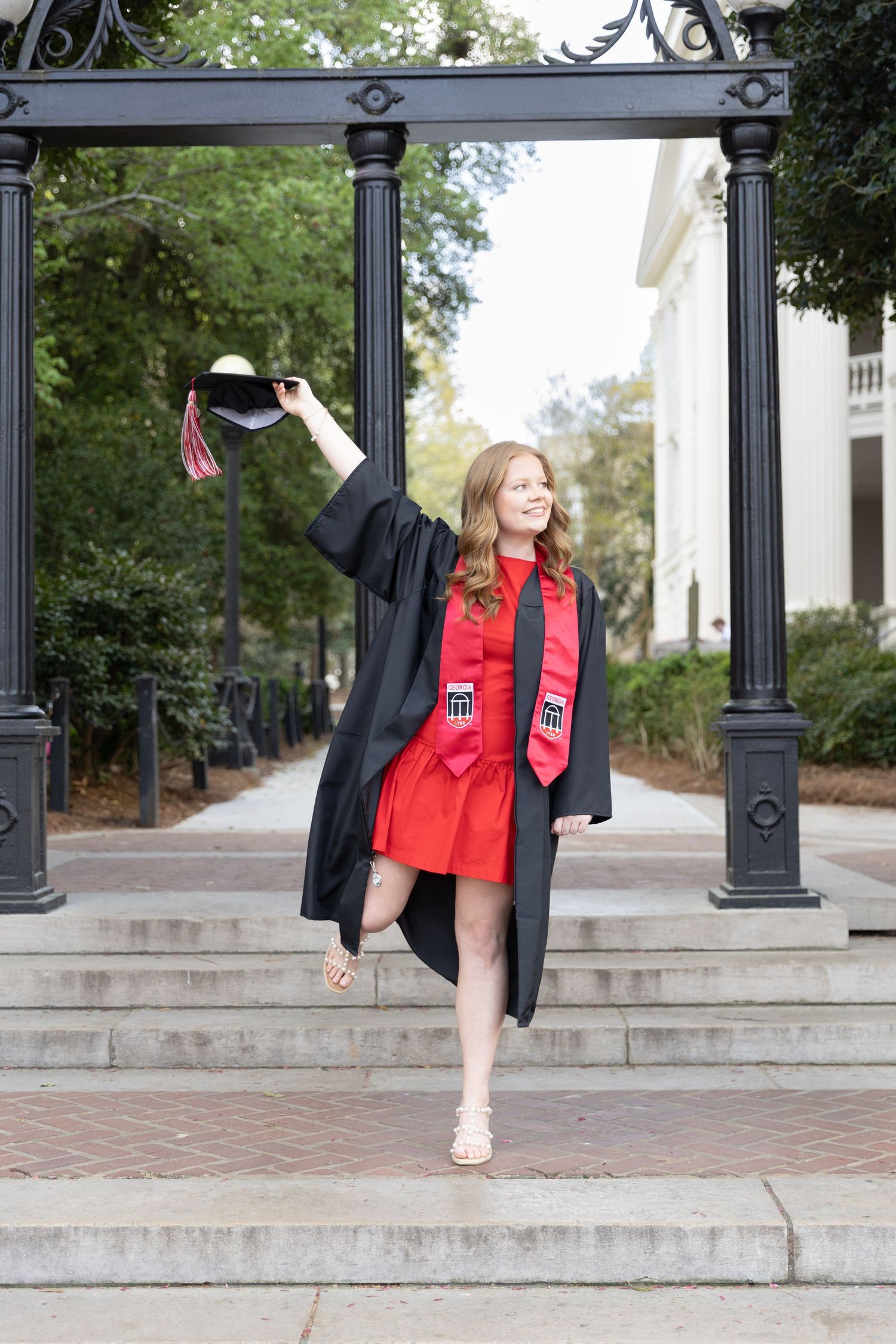 Young woman in black graduation gown and red dress raises cap triumphantly on university steps.