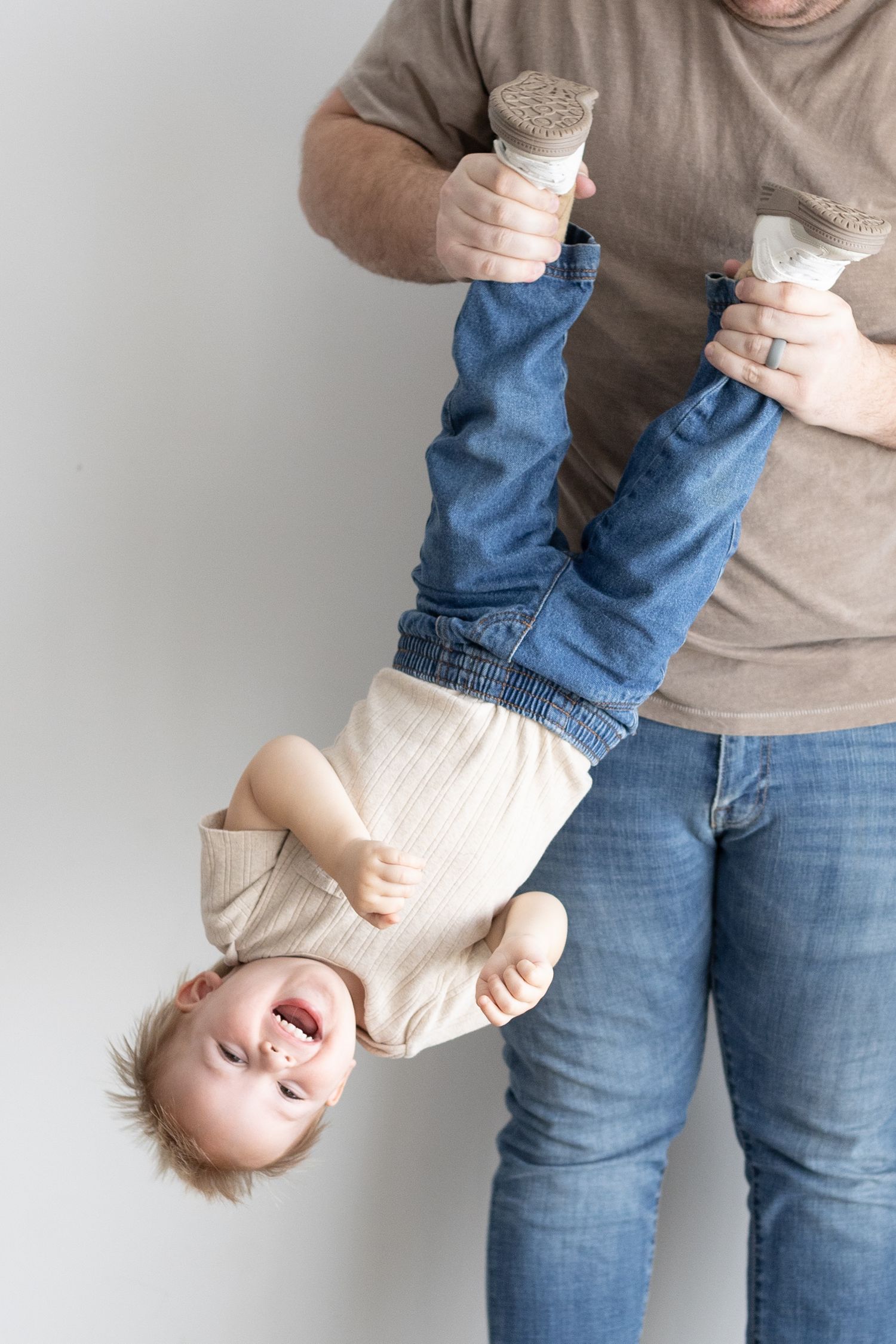 Father playfully holding laughing toddler upside down by feet, baby wearing cream onesie and blue jeans.