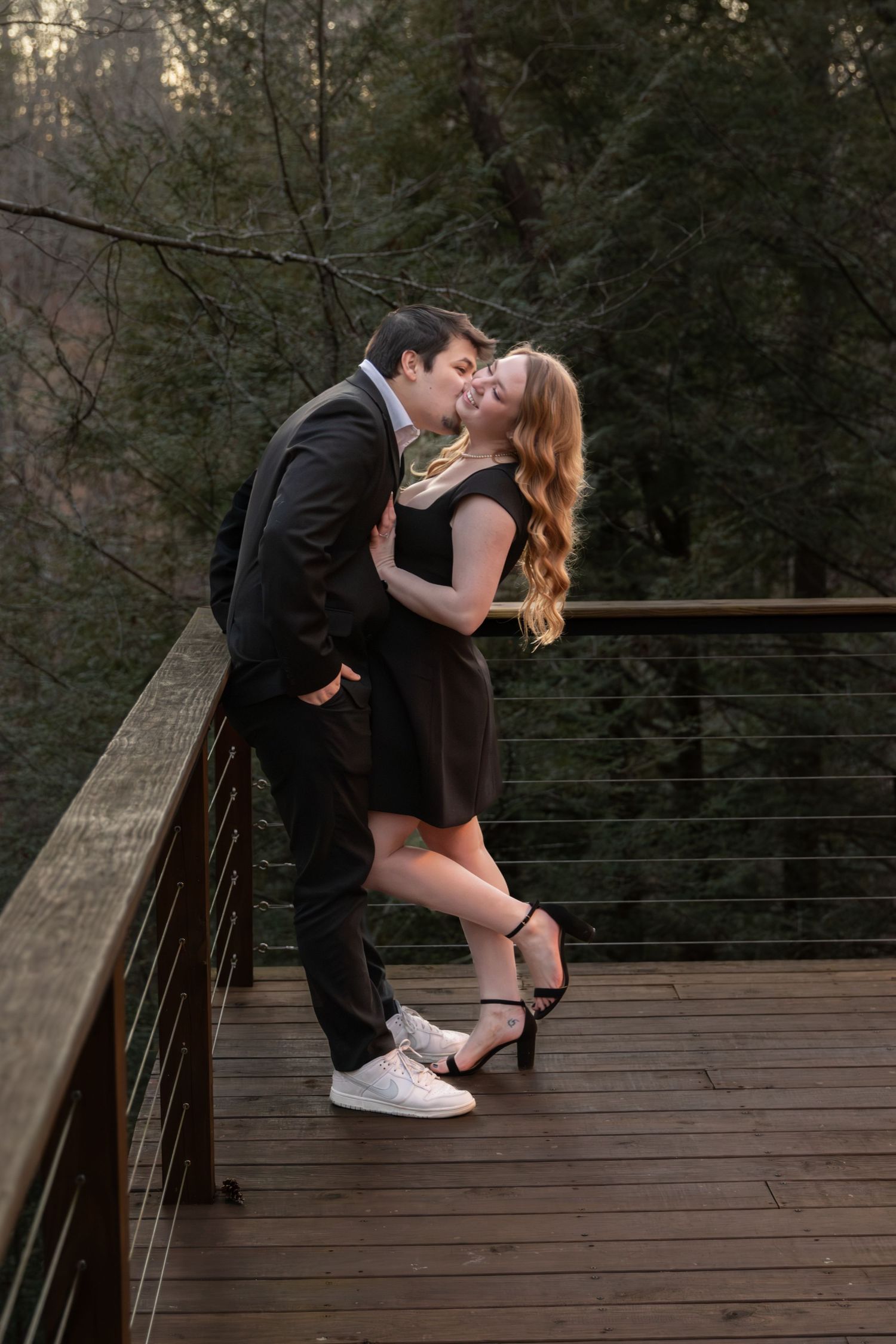 Couple sharing a romantic kiss on a wooden bridge deck surrounded by lush green trees.