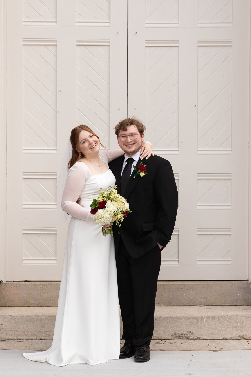Bride and groom smiling together in front of white doors, bride holding red and white bouquet in wedding attire.