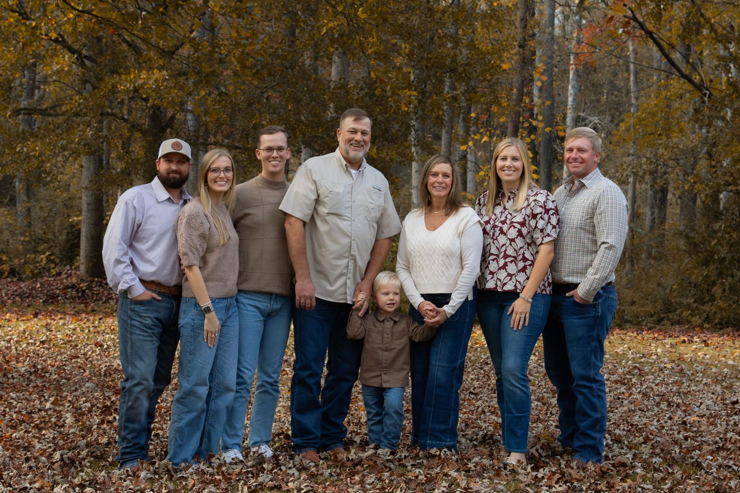 Family of eight poses outdoors in autumn forest with colorful fall foliage, wearing casual attire for a portrait.