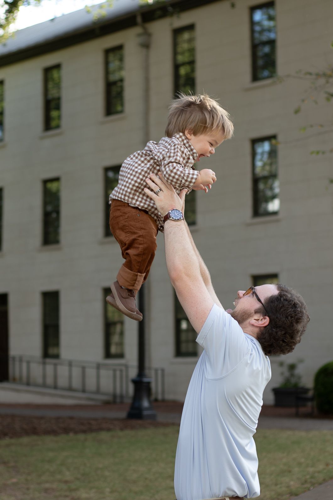 A father joyfully lifts his young son high in the air outdoors near a large brick building.