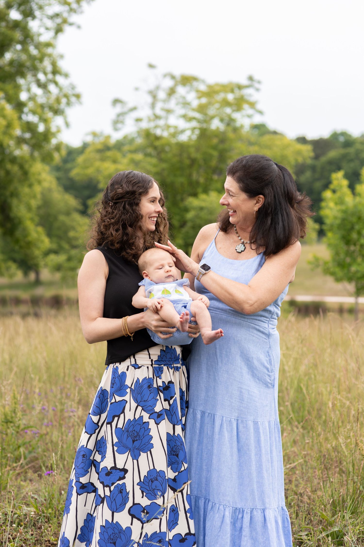 Two women smile at each other while holding a baby outdoors in a lush green field.