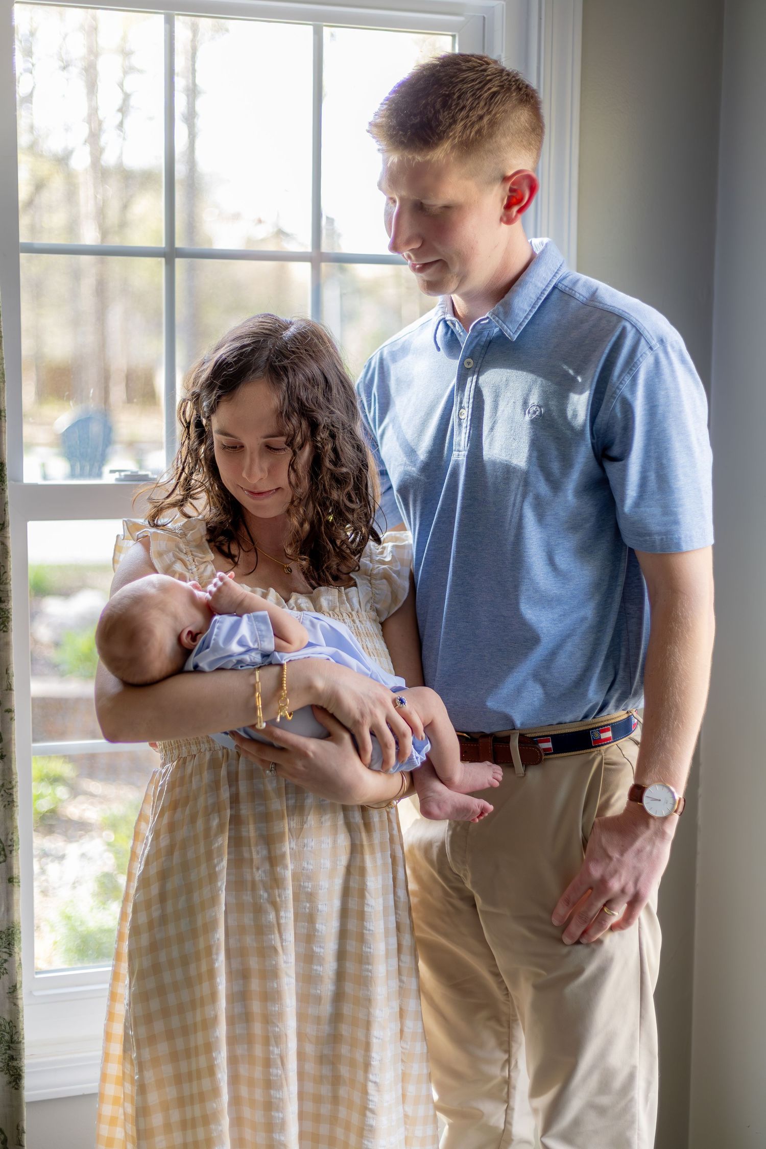 Young parents admiring their newborn baby by a sunny window, mother in yellow dress, father in blue polo shirt.