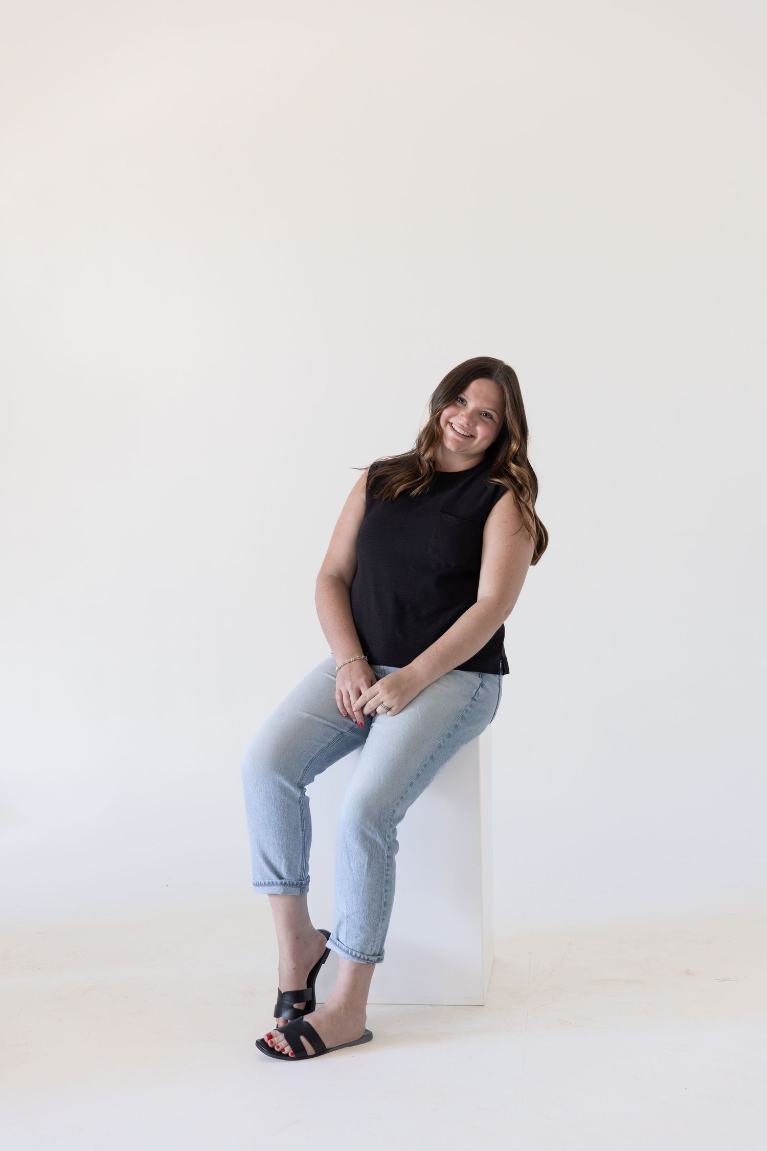 Woman in black sleeveless top and light jeans sitting on white cube, smiling against white background.