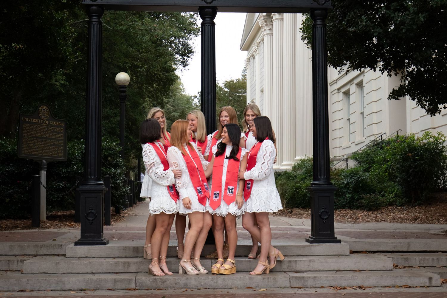 Five young women in matching white outfits with red sashes pose on steps near classic columns.