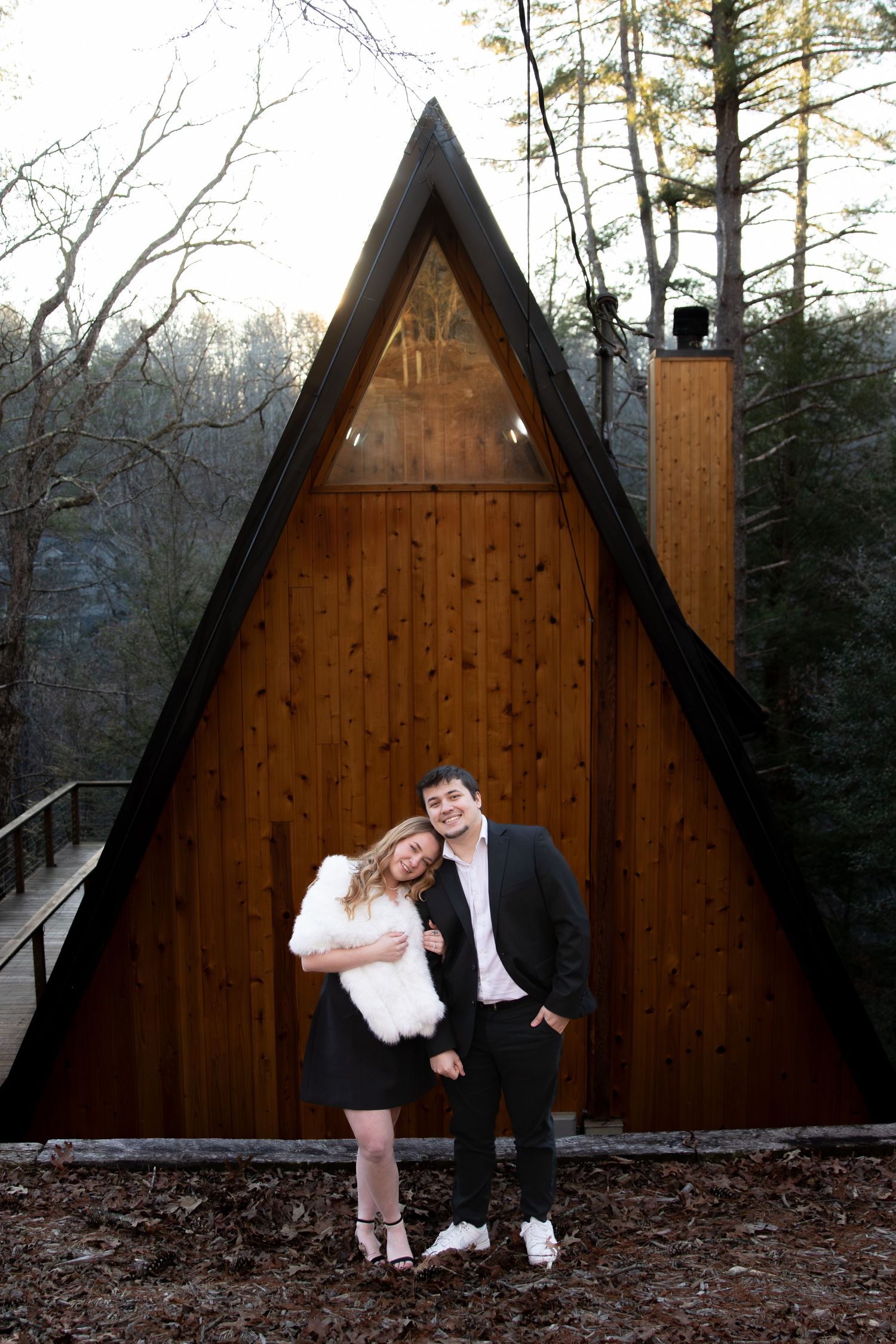 Couple posing in front of an A-frame wooden cabin in a forest setting, woman in white fur wrap and black dress.