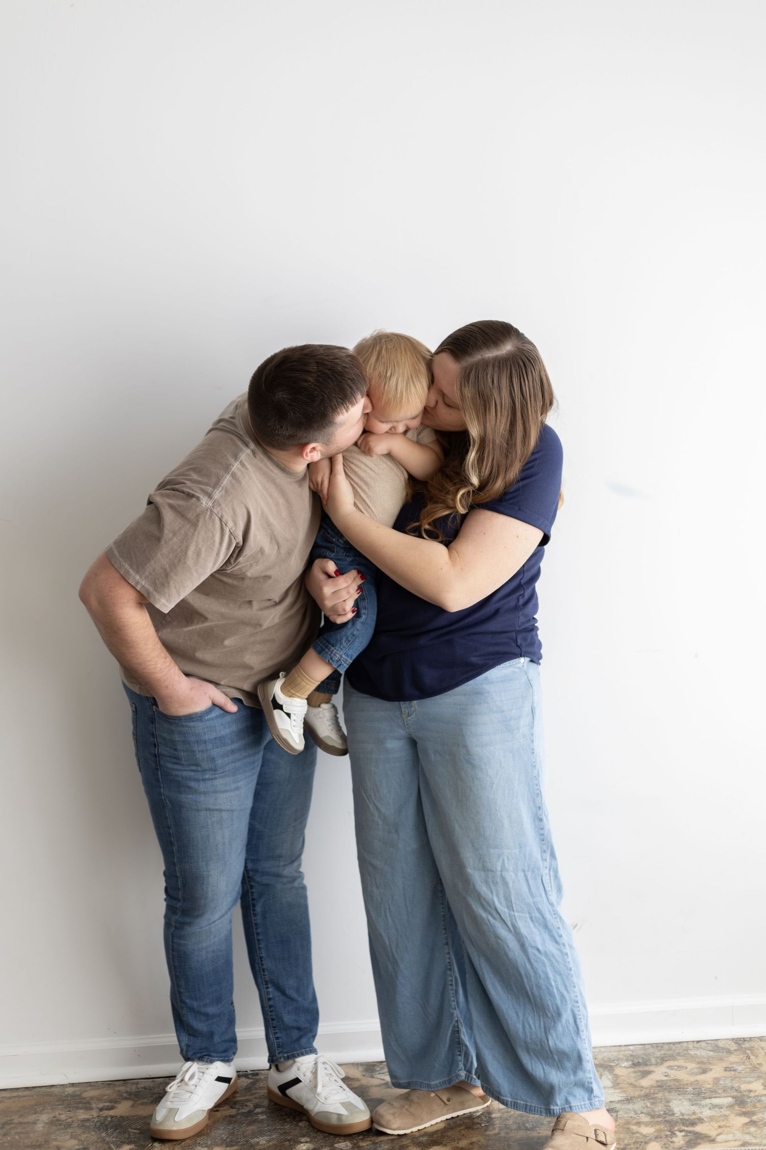 A young family shares a tender moment as two parents kiss their toddler between them against a white wall.