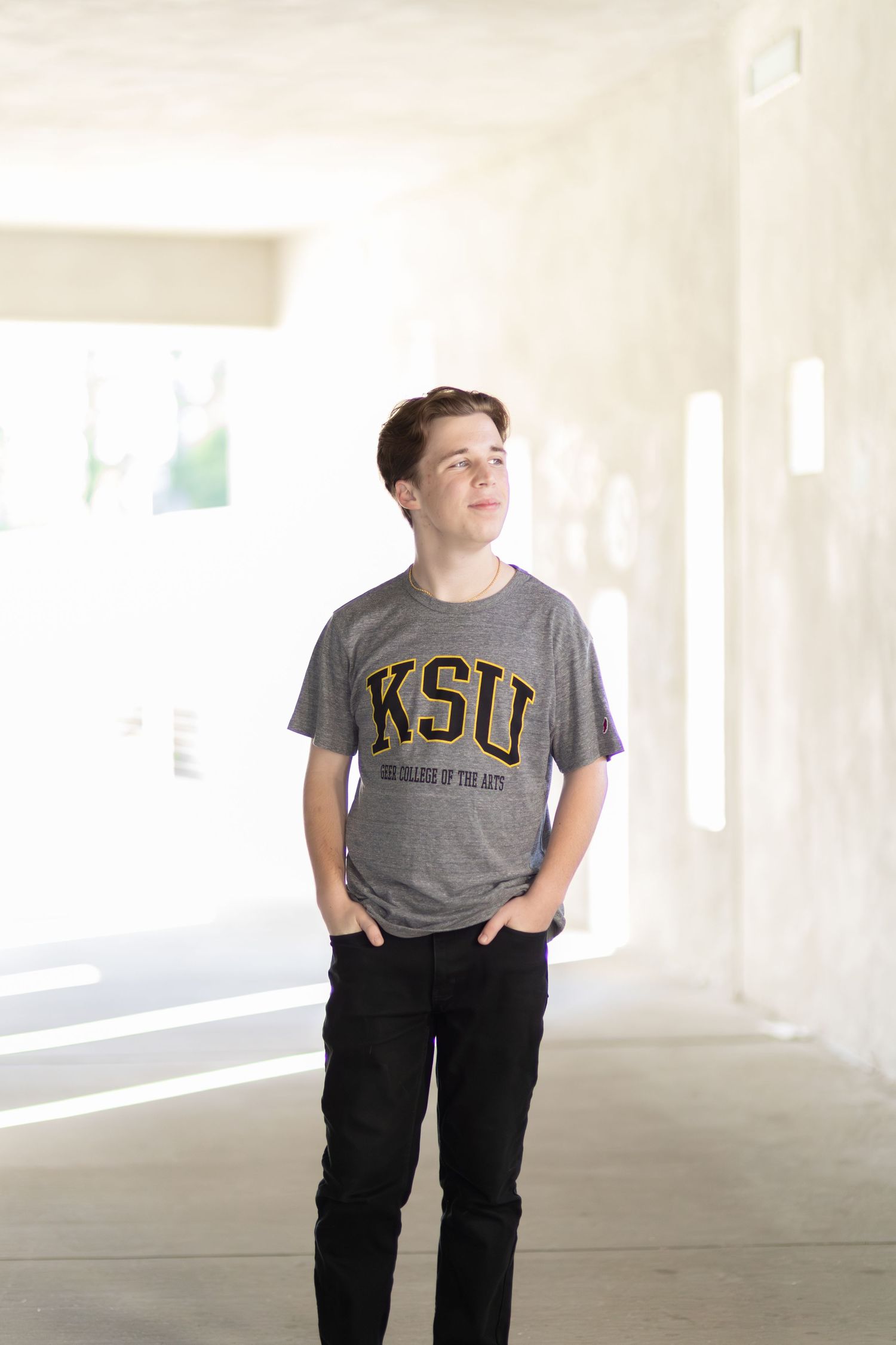Young man in KSU College of the Arts gray t-shirt and black jeans standing in a bright modern hallway.