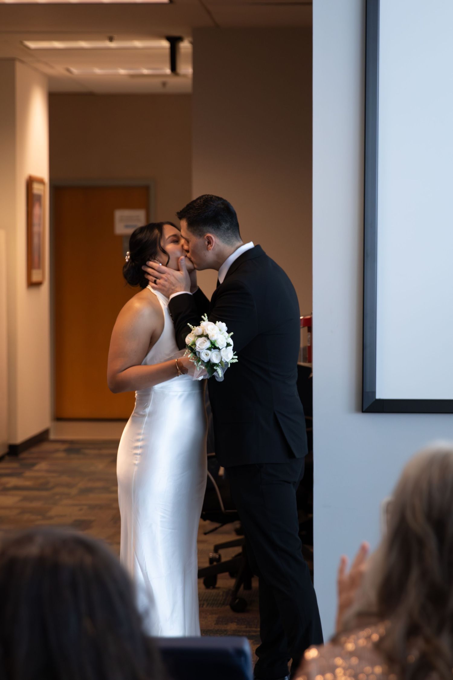Bride and groom share their first kiss in a civil ceremony, bride holding white bouquet in elegant white gown.