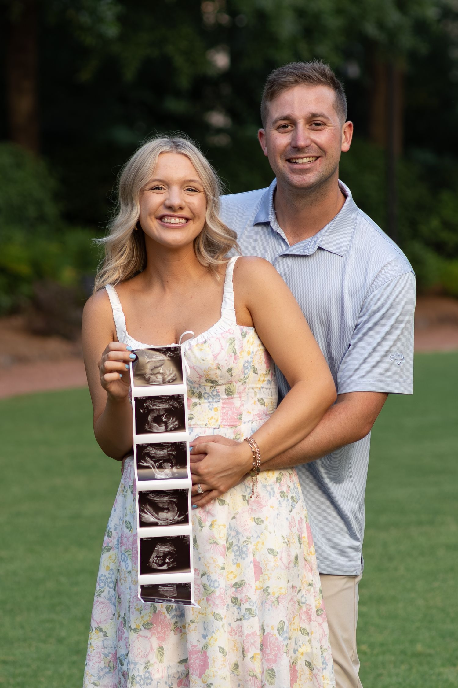 Happy couple embraces outdoors holding ultrasound strip for pregnancy reveal photo.