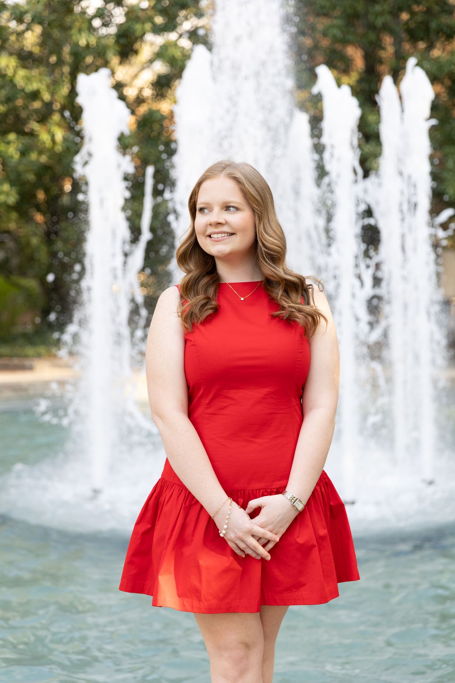 Young woman in red dress smiling in front of fountain outdoors.
