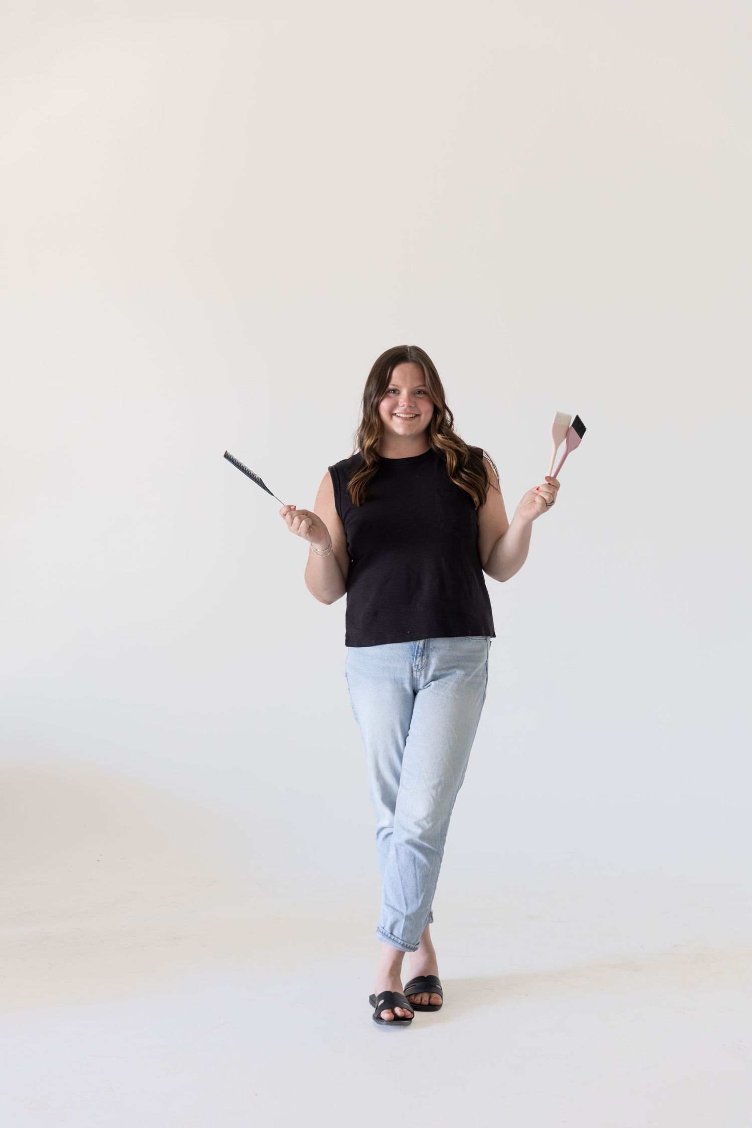 Smiling woman in black tank top and light jeans holding cooking spatulas on white background.