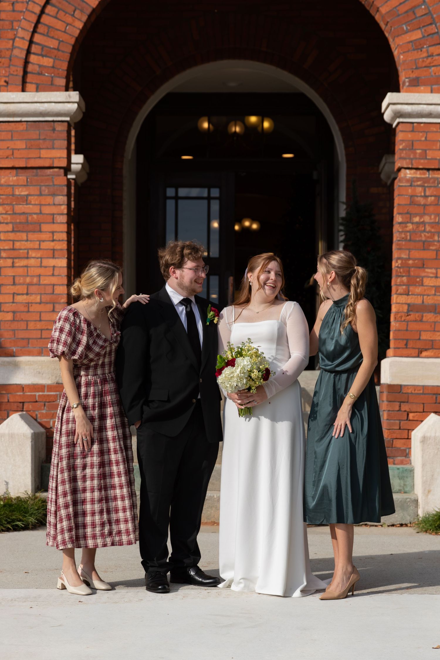 Bride in white gown with groom in black suit posing with two bridesmaids outside a red brick building.