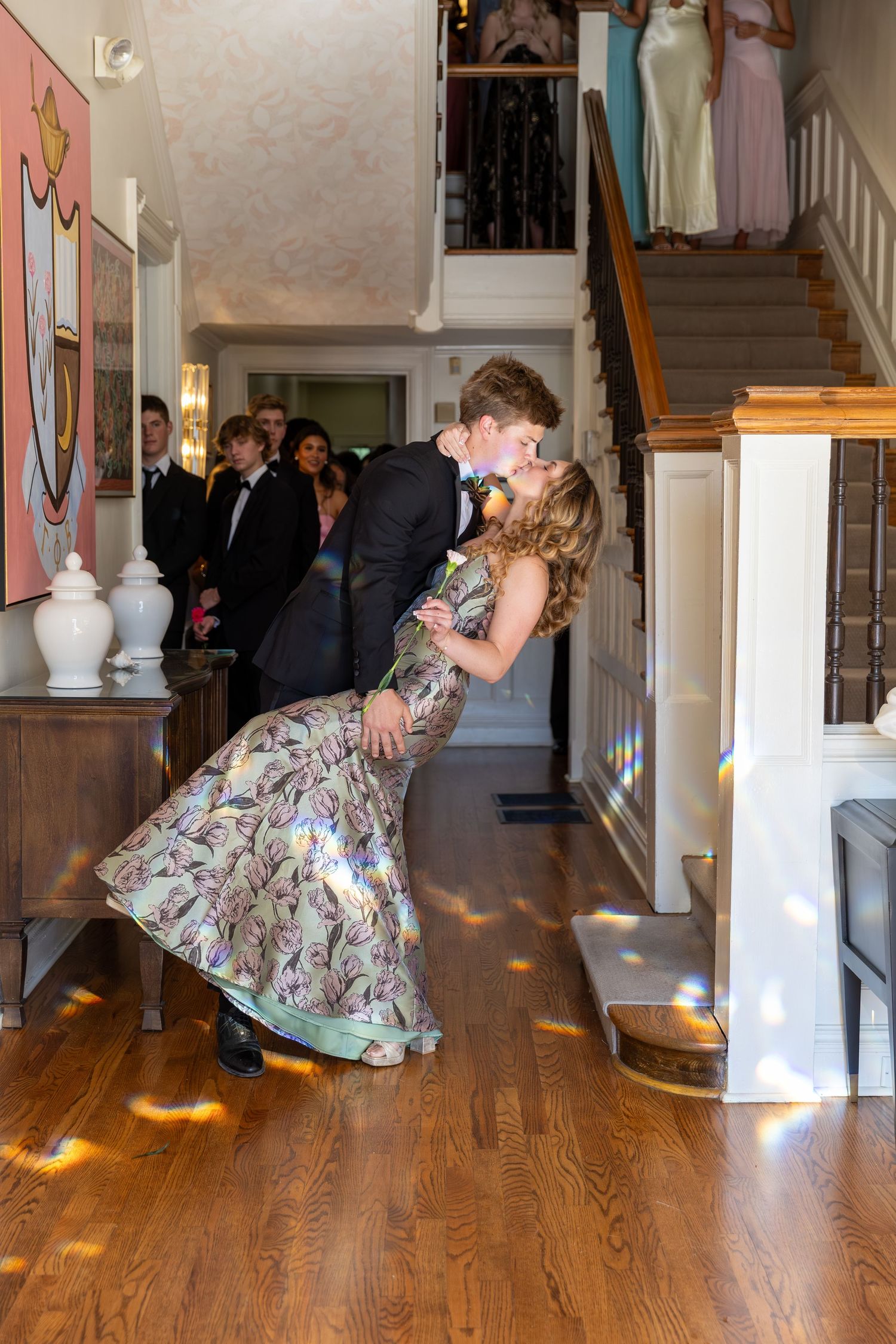 A young couple shares a romantic dip kiss in a elegant home foyer while prom guests watch from the staircase.