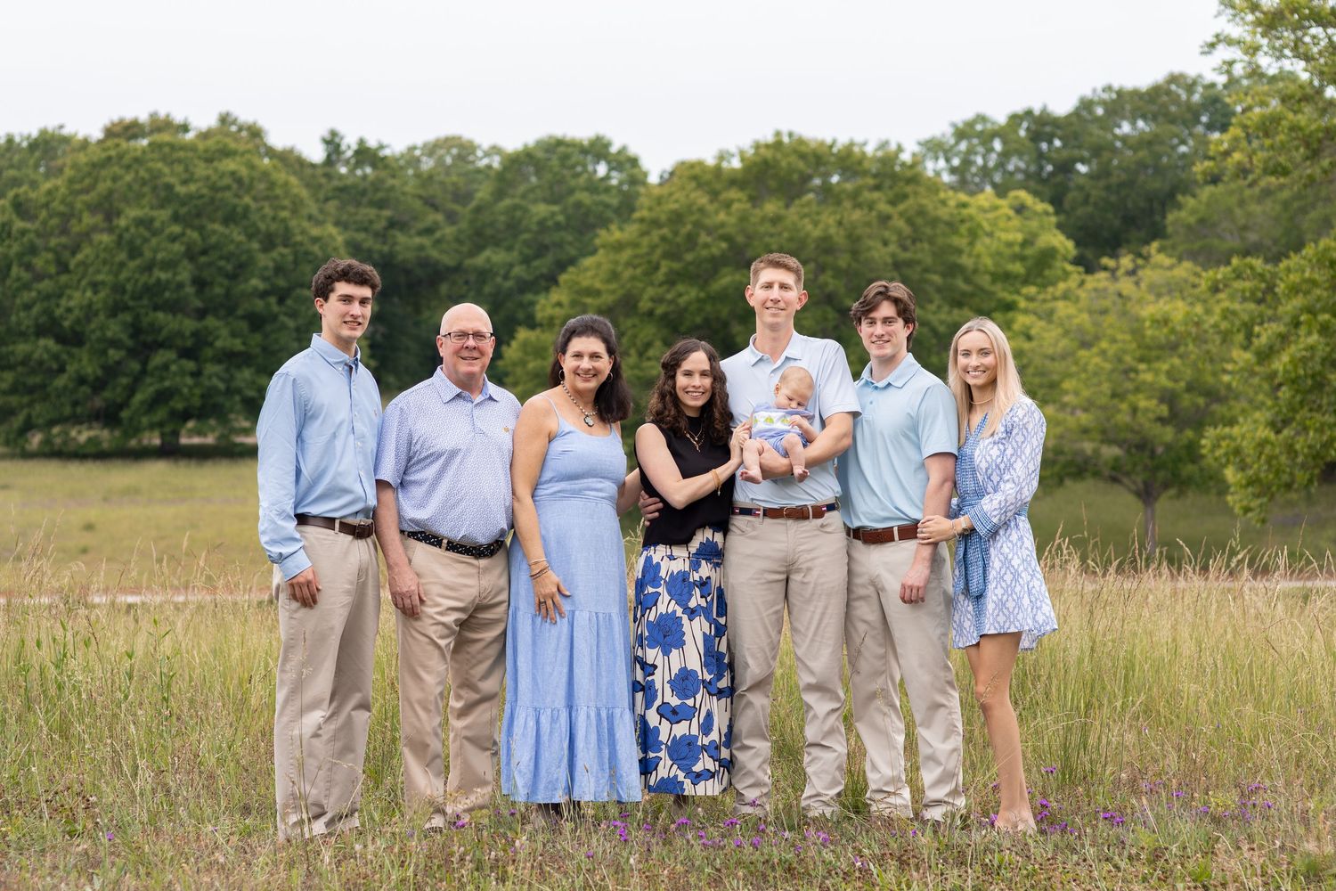 Multi-generational family of seven posing outdoors in a grassy field, including a newborn baby, dressed in blue and neutral tones.