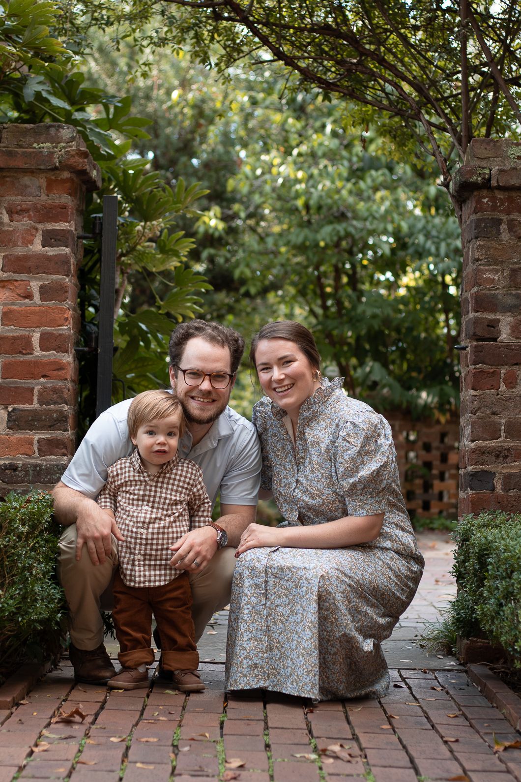 A smiling family of three poses on a brick path by a garden gate, with parents crouching beside their toddler son.