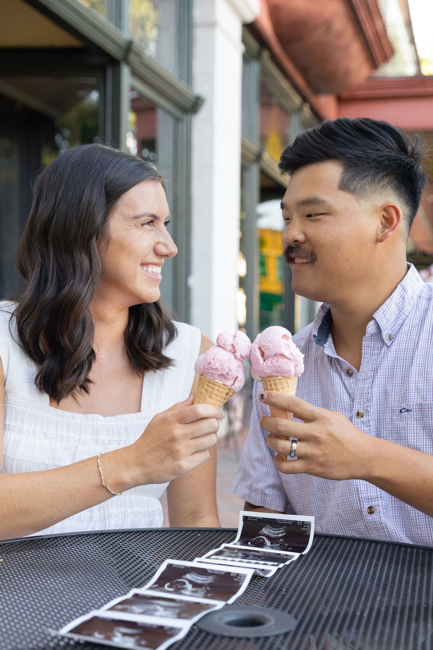 Smiling couple holding pink ice cream cones at outdoor table with ultrasound photos, celebrating pregnancy announcement.