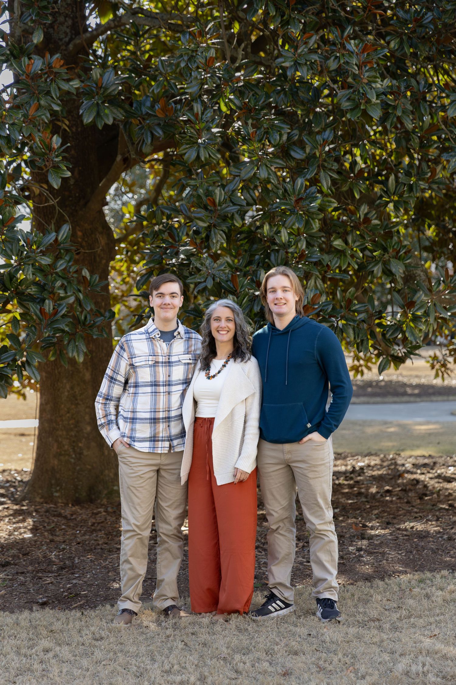 A woman and two teenage boys smile together outdoors under a large magnolia tree in coordinated fall outfits.