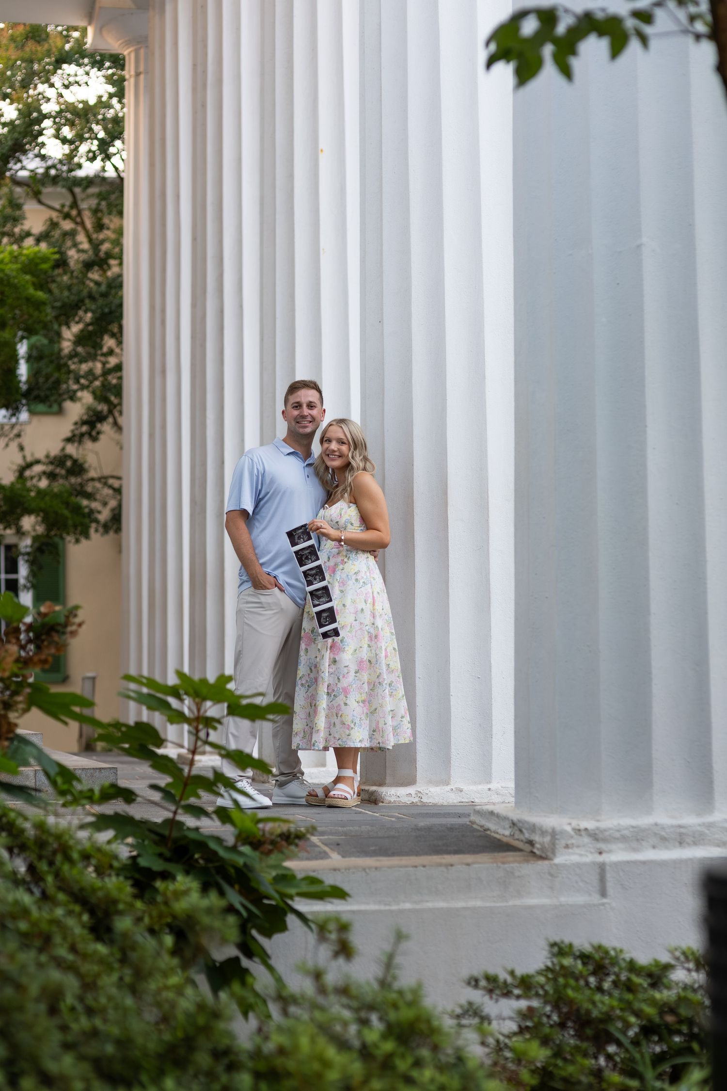 Couple holding ultrasound photos on steps of building with white columns for pregnancy announcement.