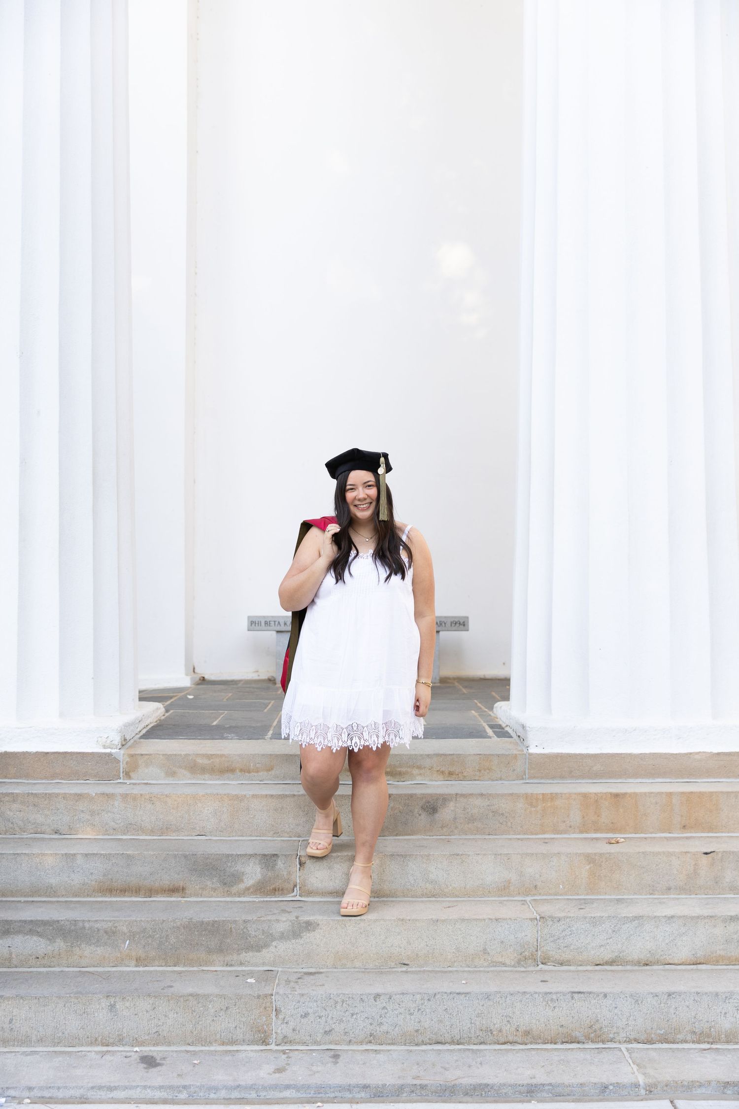 Smiling graduate in white lace dress and cap walks down stone steps between white columns.