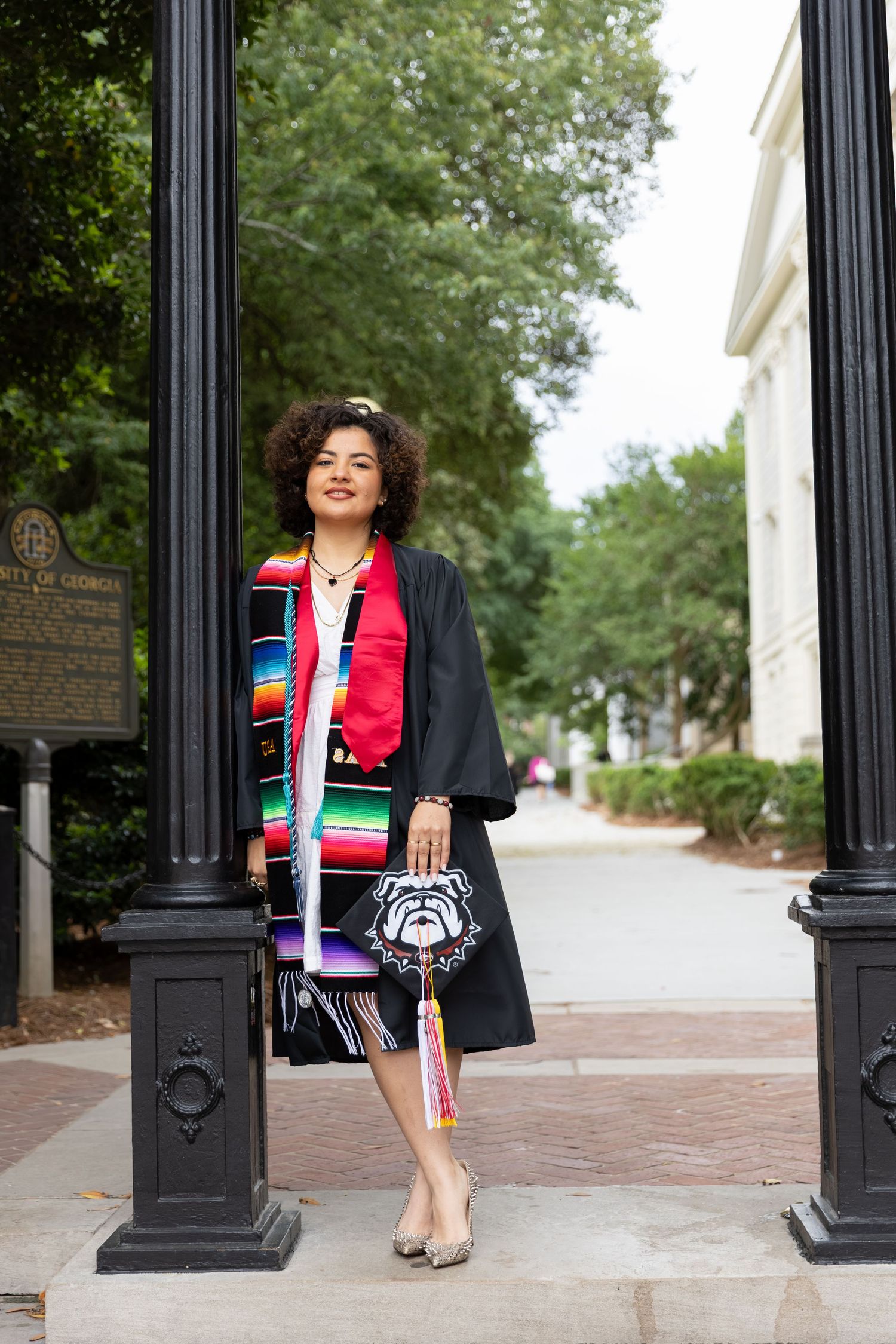Graduate in black gown with colorful stole holds decorated UGA bulldog cap between campus pillars.