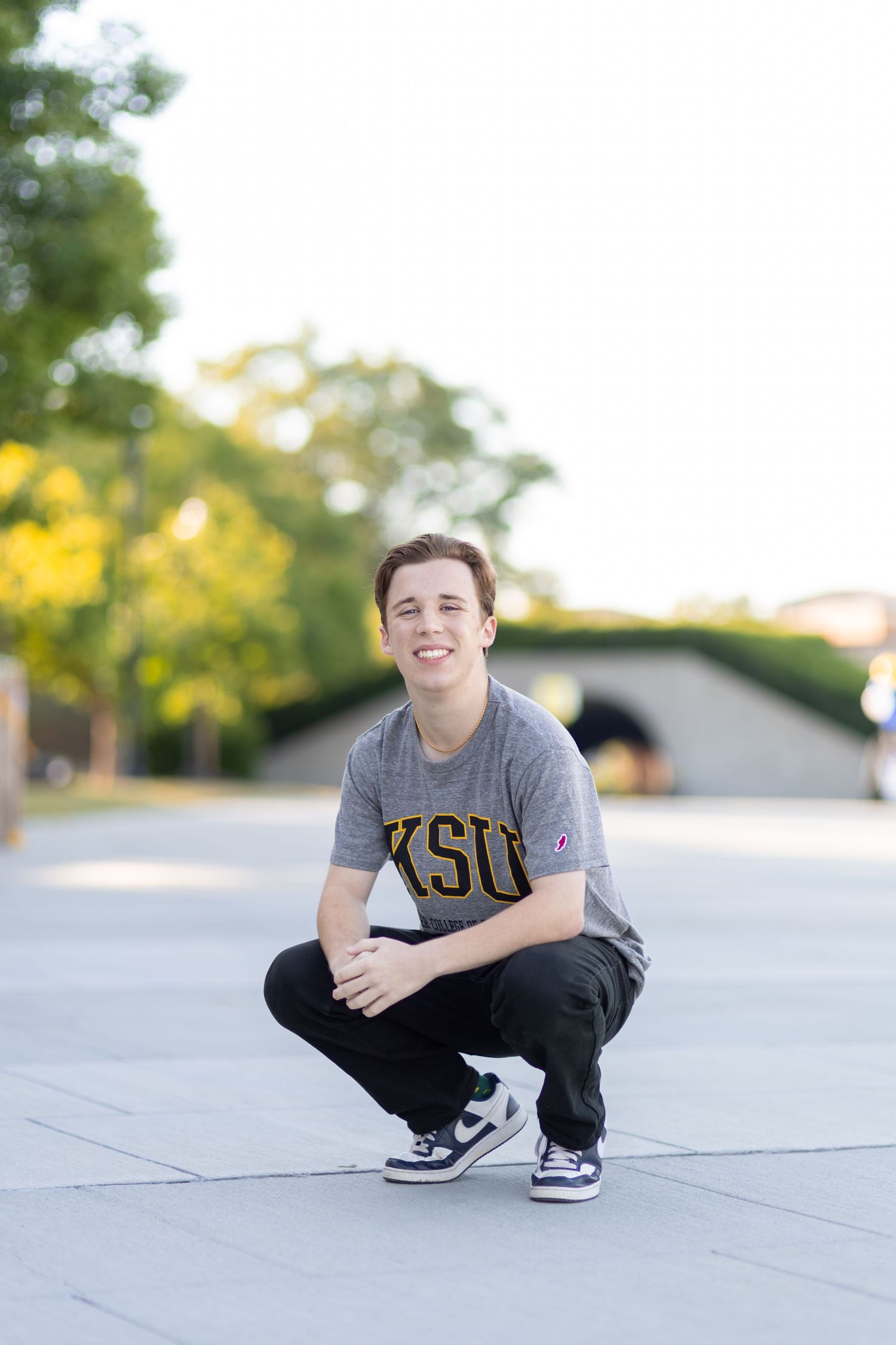 Young man in KSU gray t-shirt crouching and smiling outdoors on a sunny day.