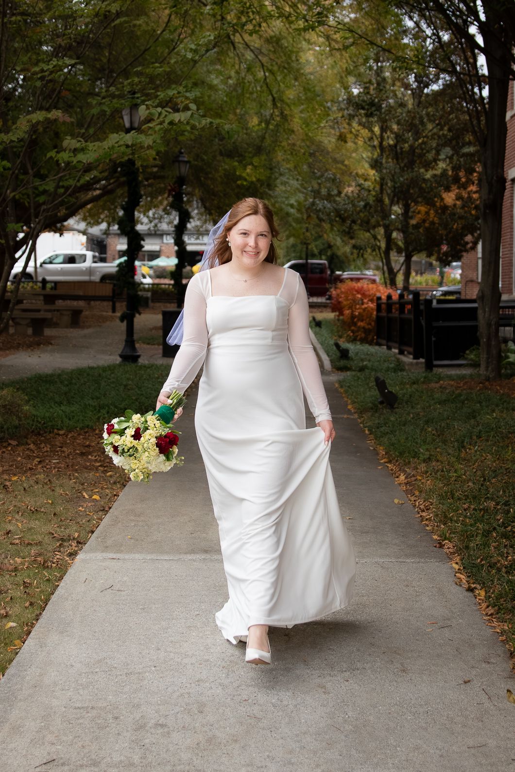 Smiling bride in white long-sleeve gown walks along outdoor sidewalk holding colorful bouquet with trees in background.