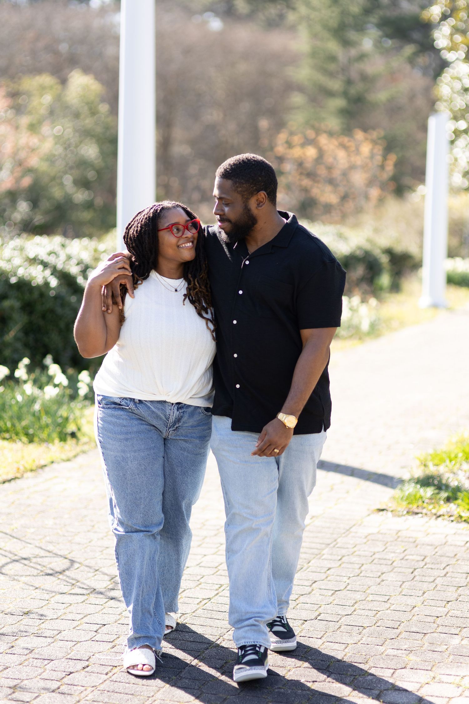 A happy Black couple walks arm in arm on a sunny brick path, smiling at each other in casual jeans and tops.