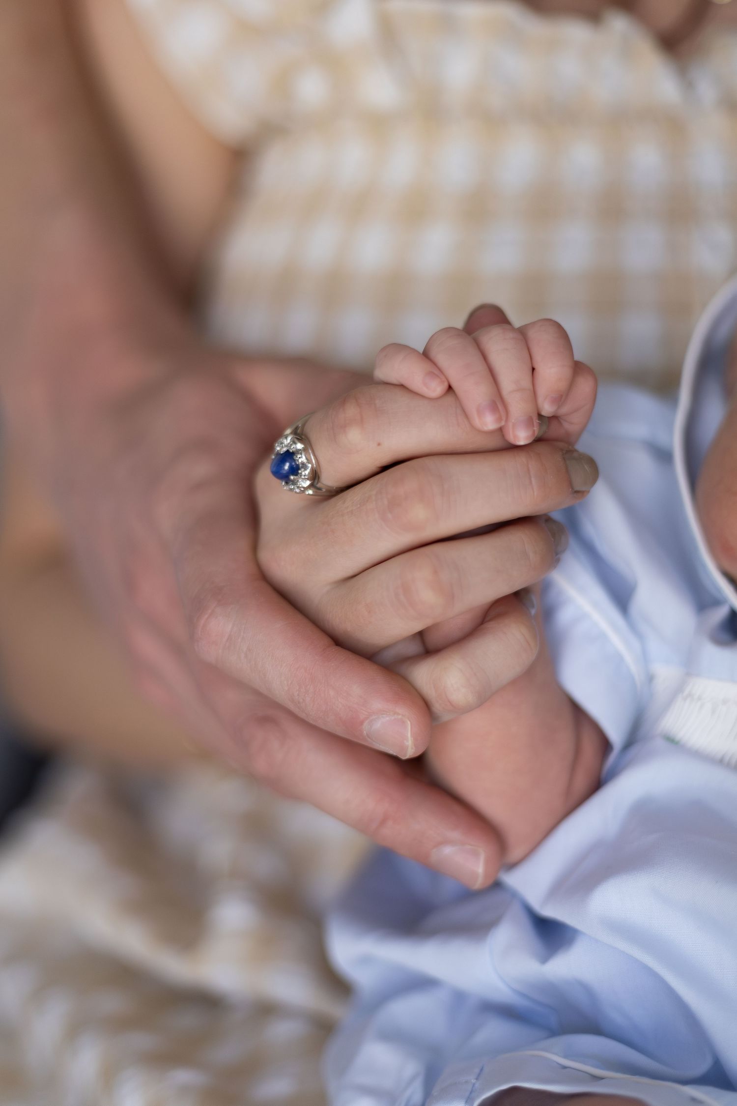 A mother's hand with a blue gemstone ring gently holds her newborn baby's tiny fingers in a tender moment.