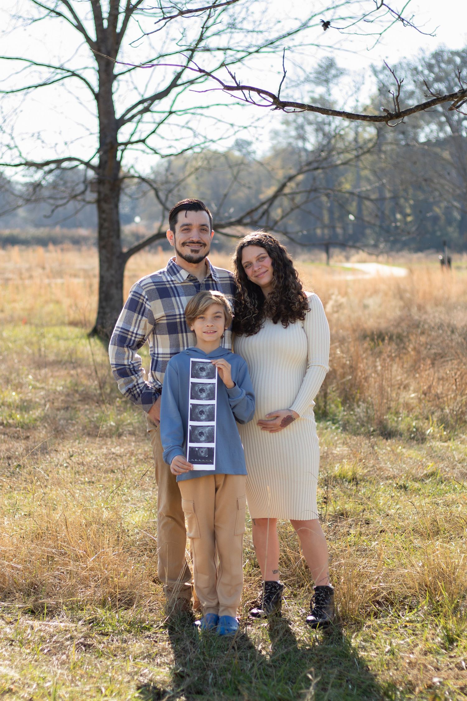 Pregnant woman in cream dress poses with husband and son holding ultrasound photos outdoors in a sunny field.