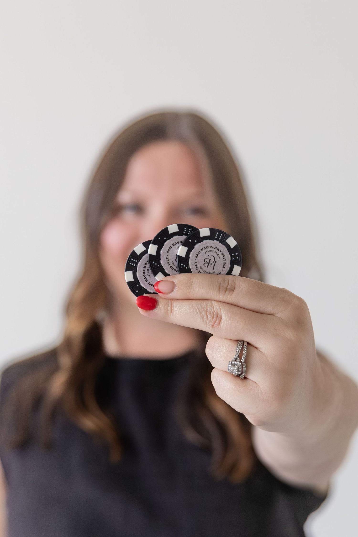 Woman in black top holding two black poker chips toward camera, wearing a diamond ring with red nail polish.