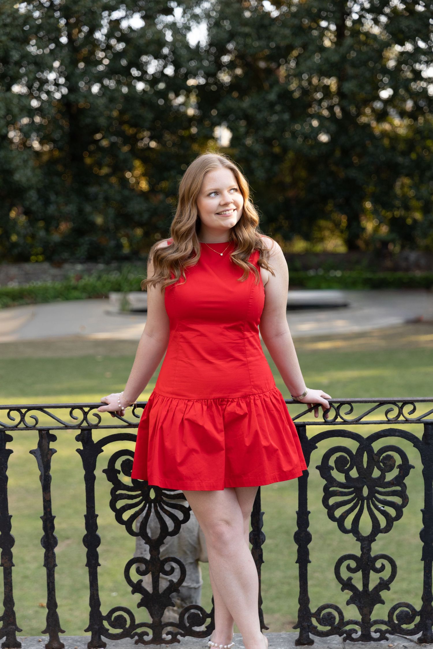 Young woman in red sleeveless dress posing by ornate iron fence in garden setting.