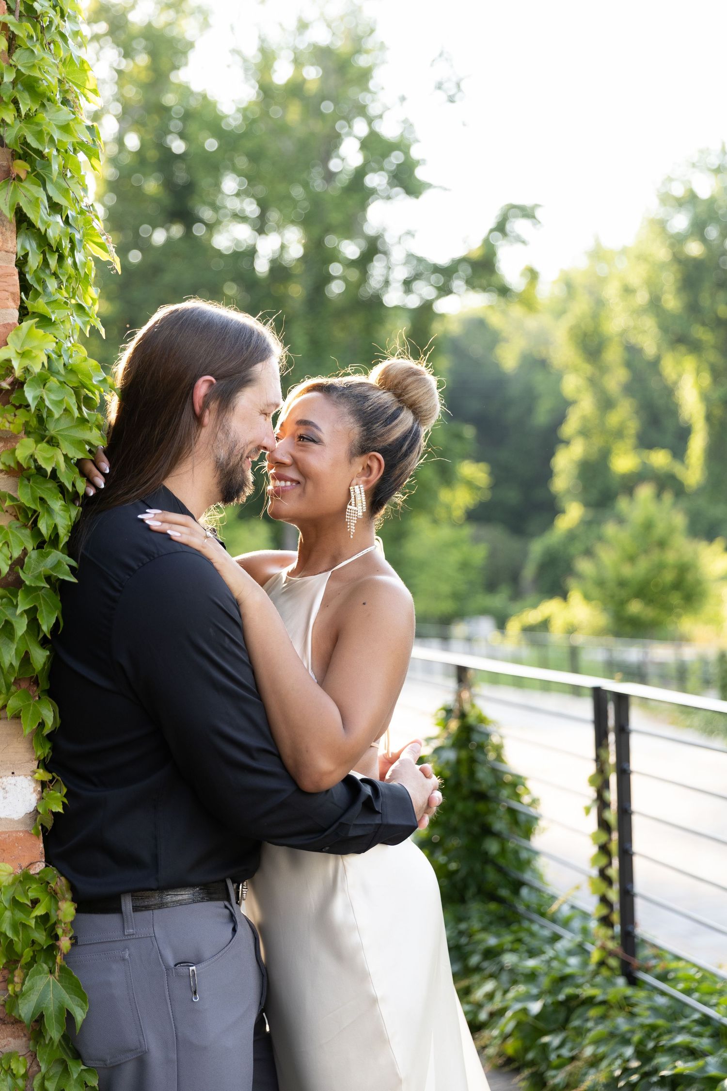 Smiling bride in white dress embraces groom in black suit on sunlit outdoor balcony surrounded by lush green ivy.