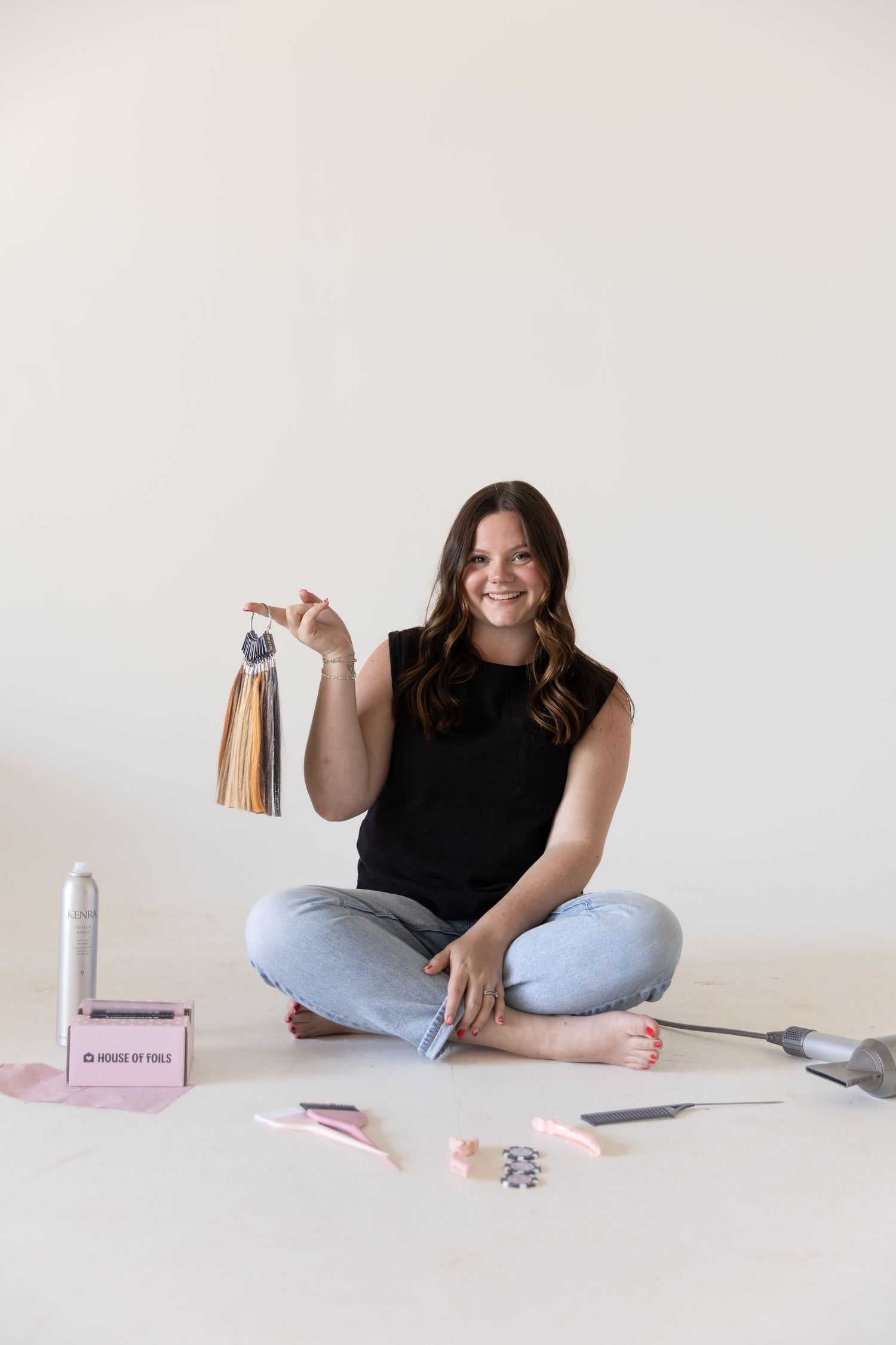 Woman sitting cross-legged on floor holding hair extensions, surrounded by styling tools and products, smiling at camera.