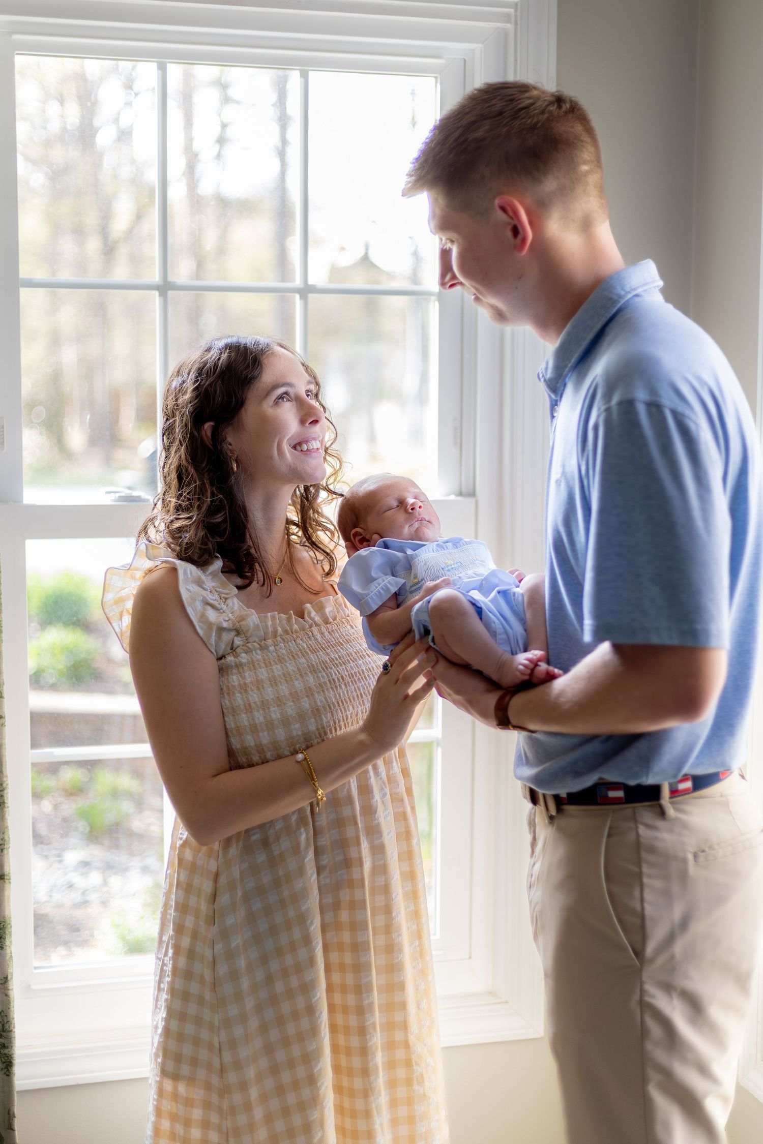 A young couple stands by a bright window, smiling as the man holds their newborn baby wrapped in a light blue blanket.