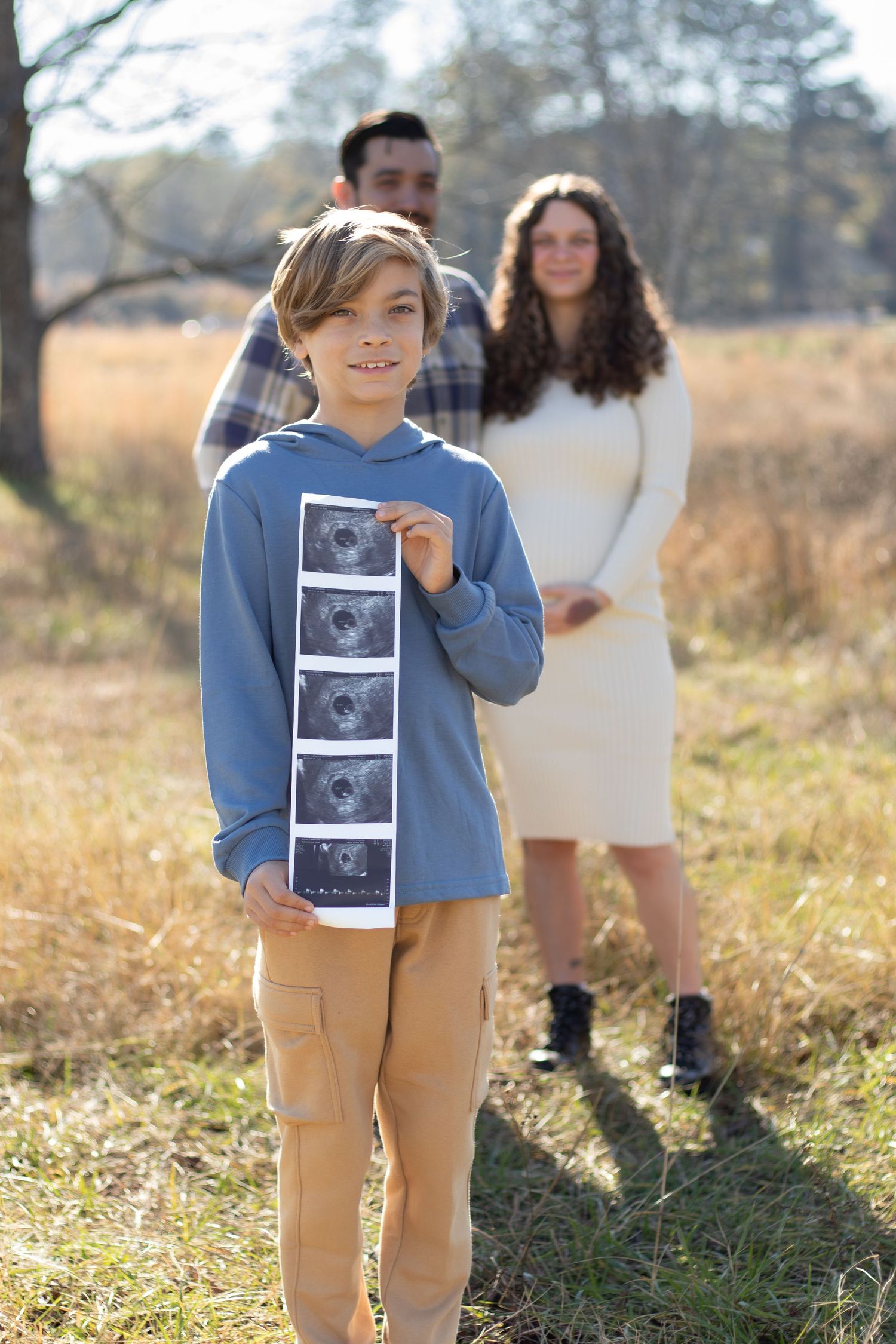 A smiling boy holds ultrasound photos outdoors as his pregnant mother and father stand behind him in a field.
