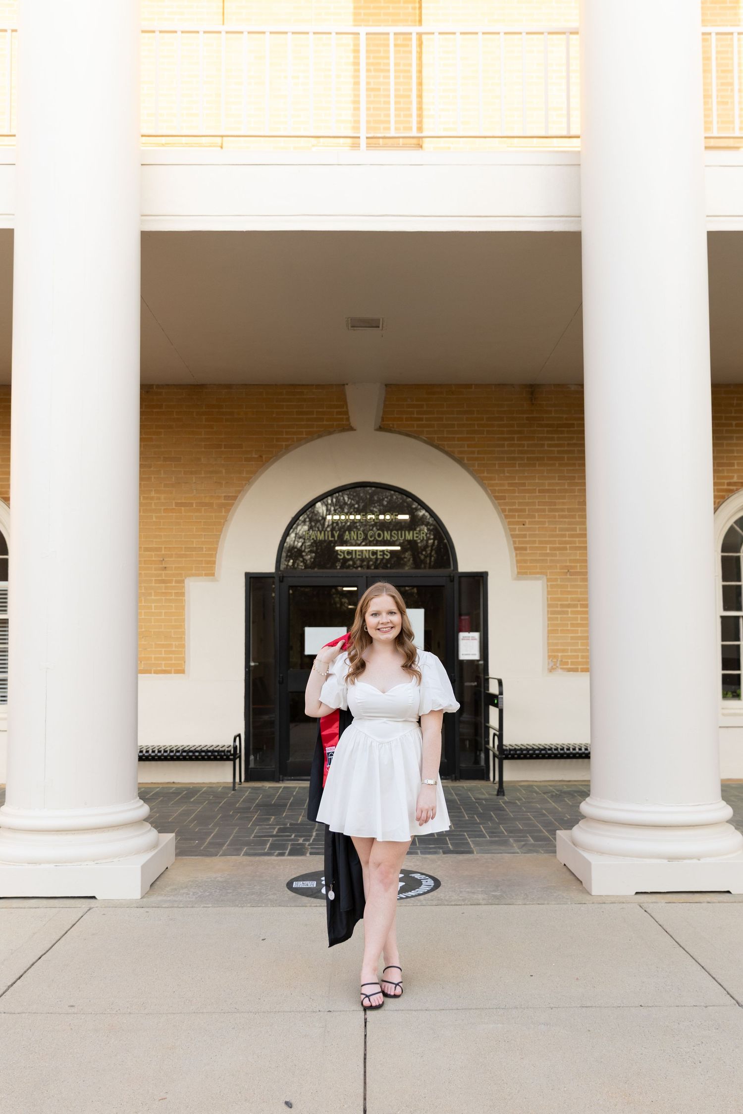 Young woman in white dress holding graduation cap outside brick building with white columns.