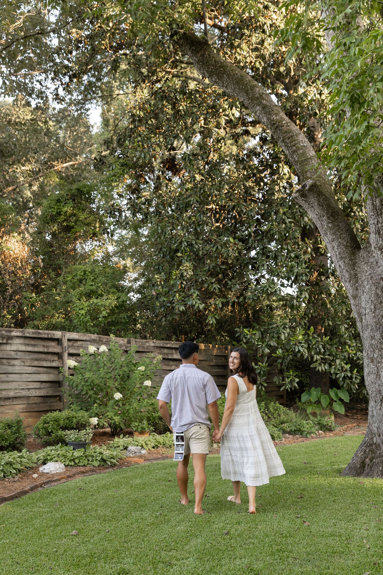 Couple holding hands walking barefoot in lush garden under large oak tree, woman in white dress looks back.