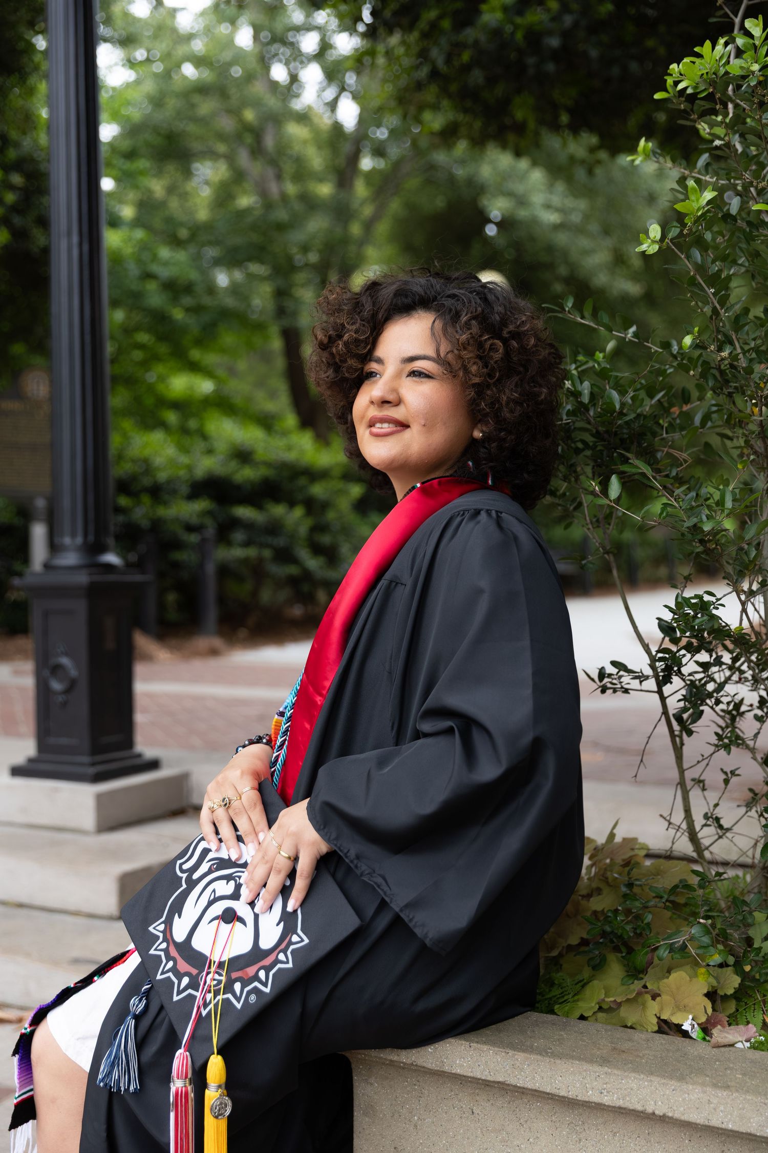 Smiling graduate in black cap and gown with red sash sits outdoors, decorated mortarboard with bulldog design.