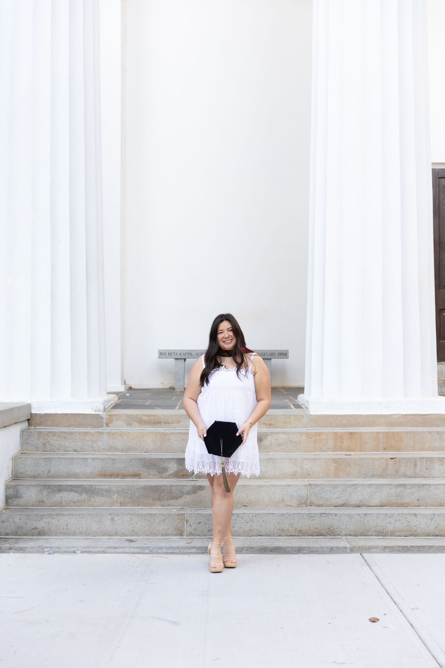 Young woman in white lace dress holding black clutch poses on stone steps between white columns.