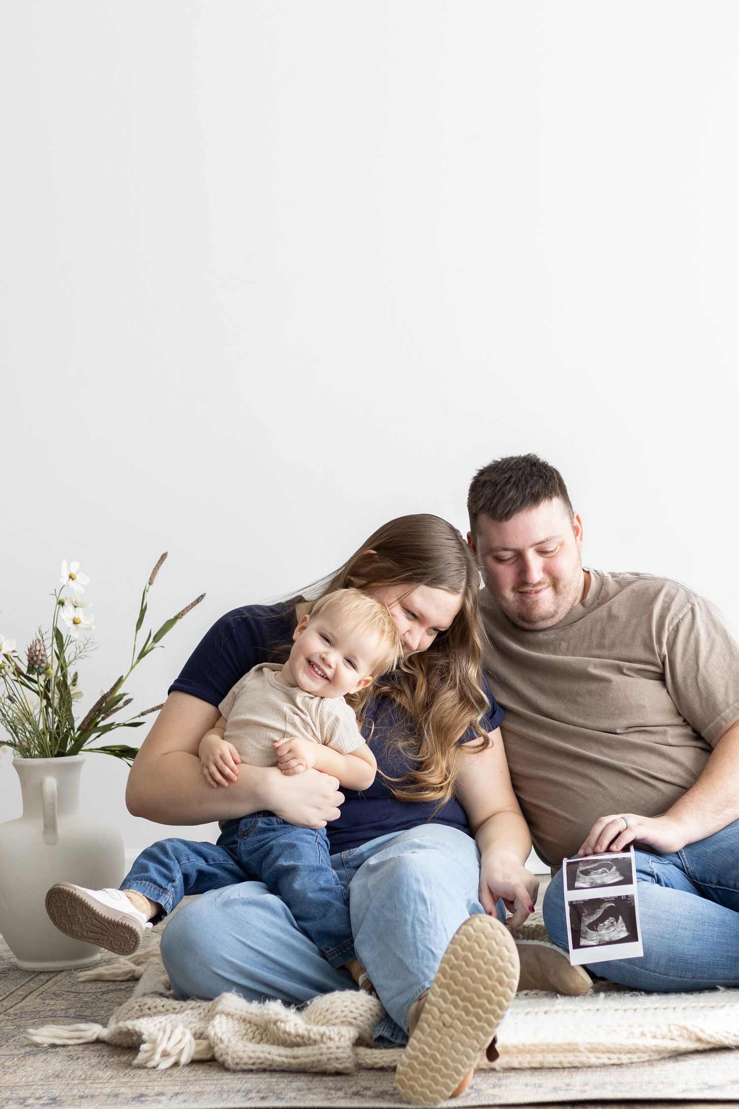 Happy couple sitting on floor with toddler, holding ultrasound photo, announcing second pregnancy at home.