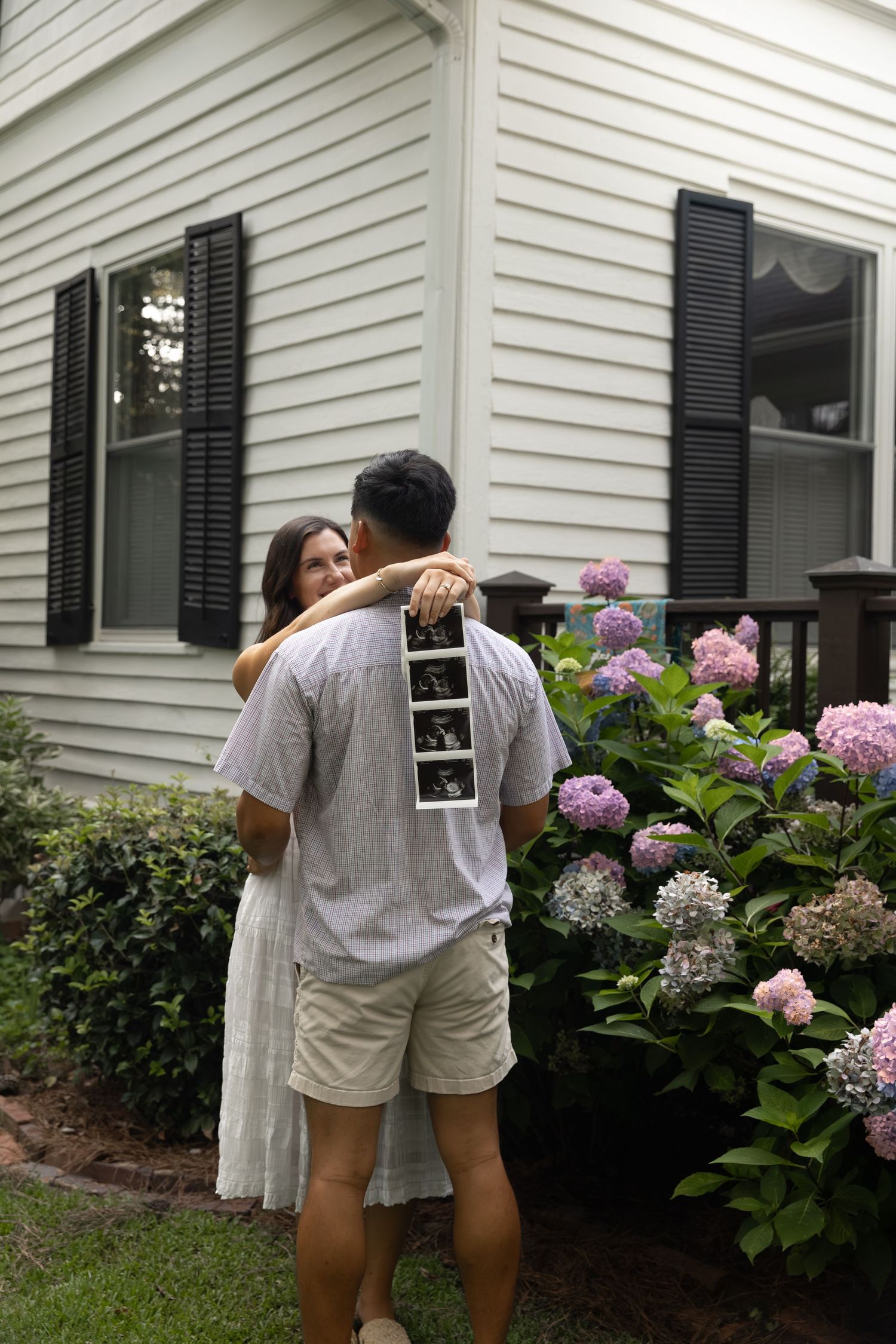 Couple embracing in garden holding ultrasound photos, announcing pregnancy outside white house with blooming hydrangeas.