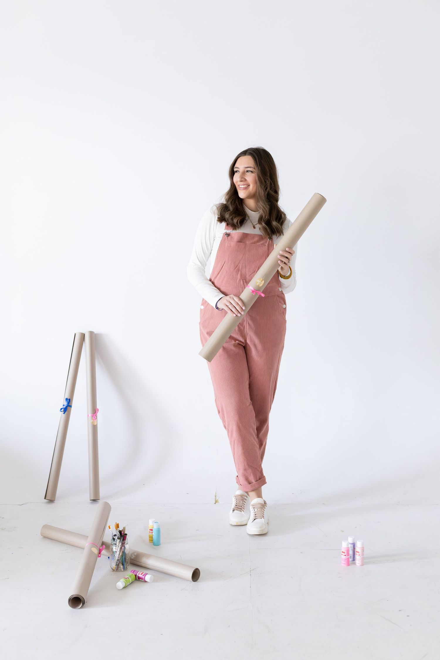 Woman in pink overalls holding rolled paper tubes surrounded by craft supplies on white background.