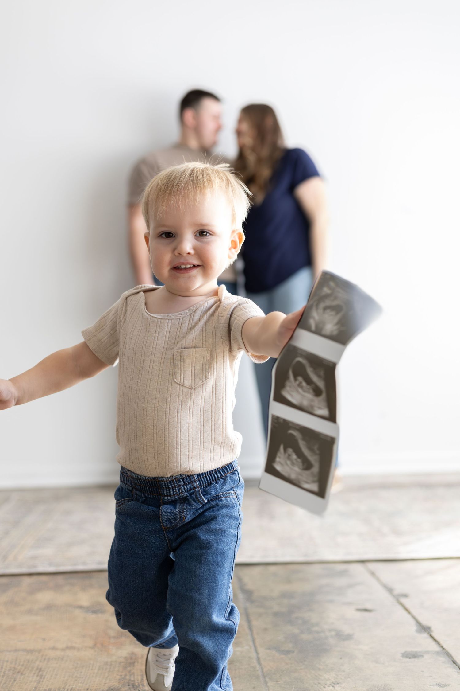 Toddler boy running and holding ultrasound photos while expecting parents stand in background.