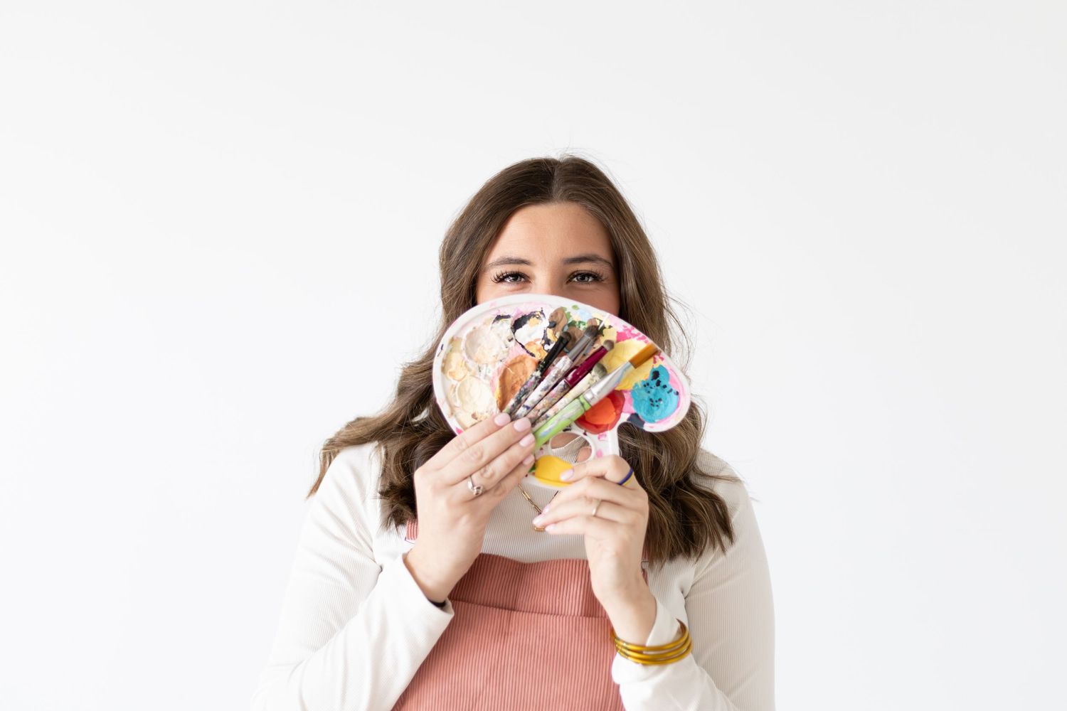 Woman holding a colorful paint palette and brushes in front of her face against a white background.