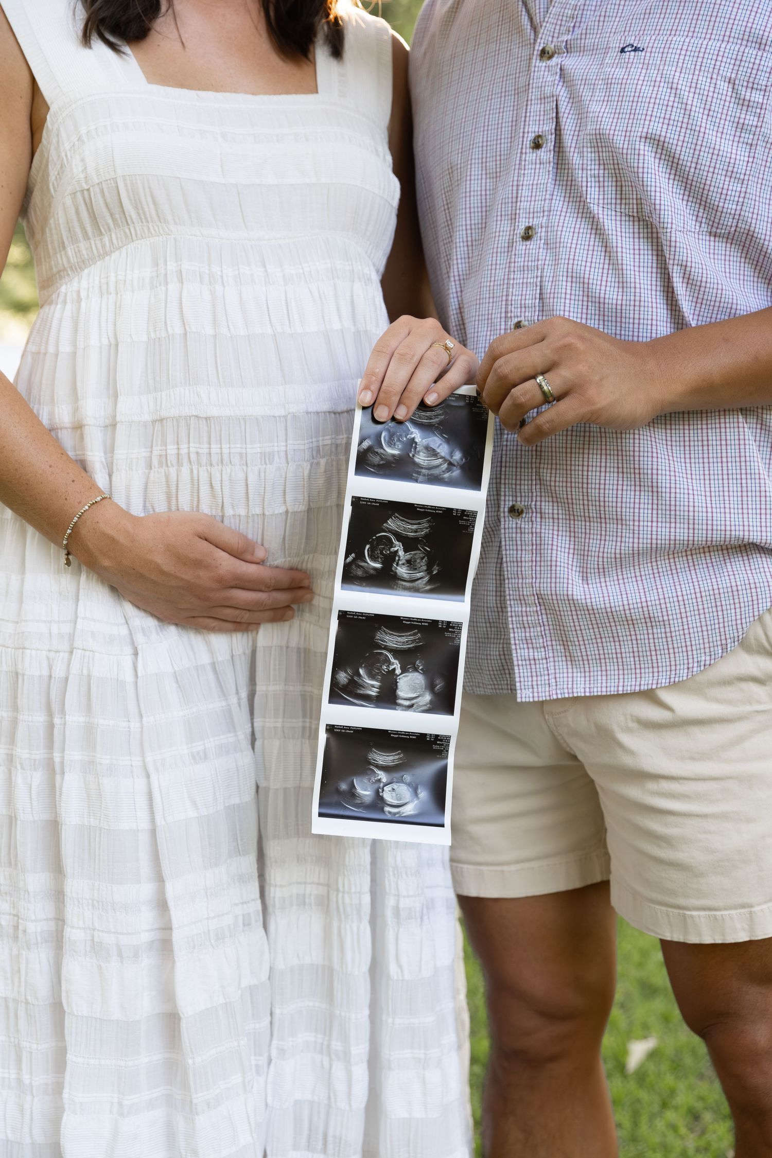 Pregnant woman in white dress and man in plaid shirt holding ultrasound photos outdoors.