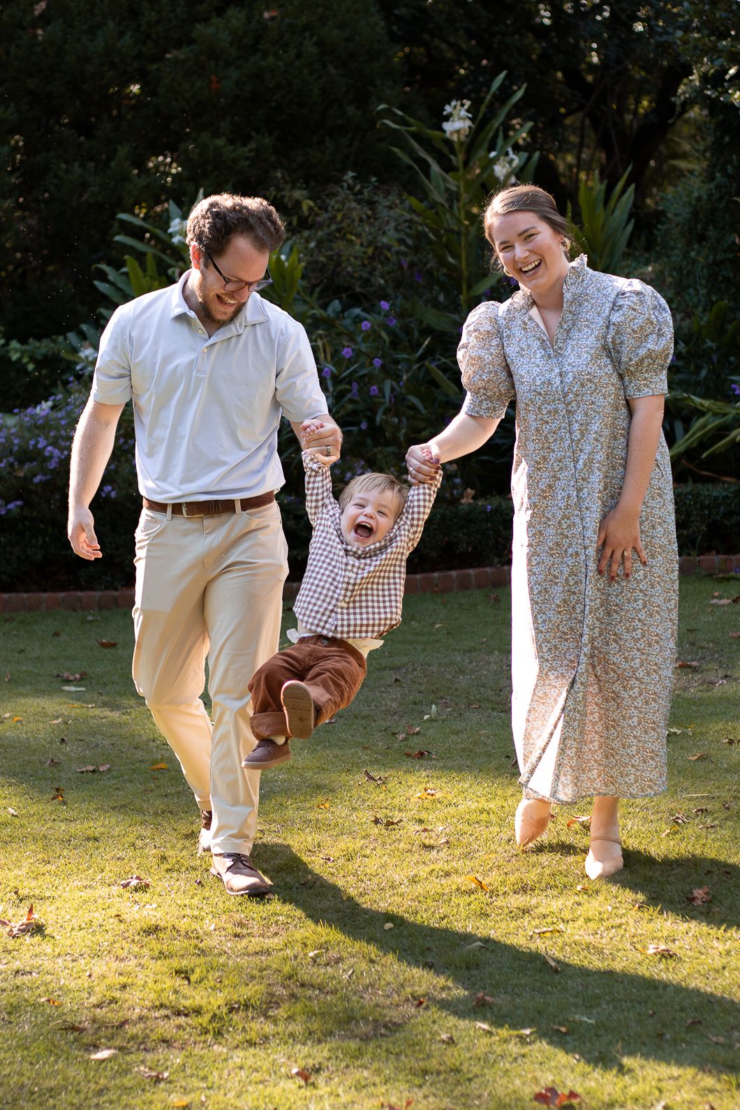A joyful family swings their laughing toddler between them while walking on green grass in a sunny garden.