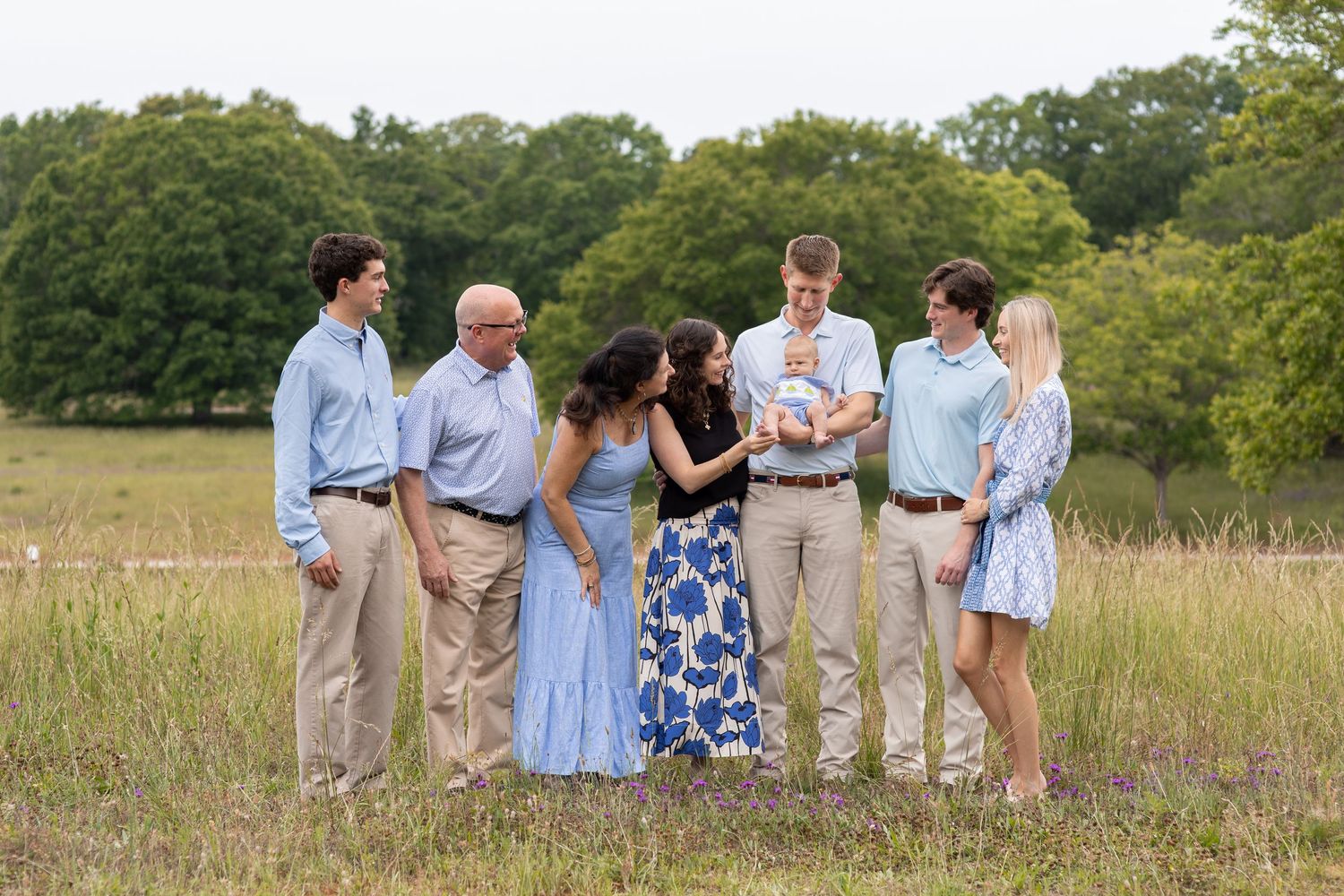 Family of six adults gathered in a sunny meadow, admiring a newborn baby held by a tall young man.